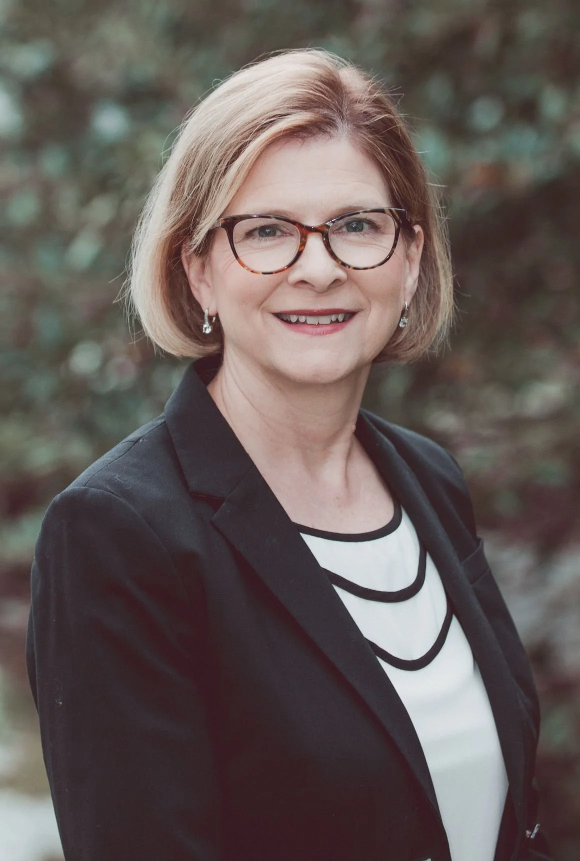 A woman with short blonde hair, glasses, and earrings, wearing a black blazer and a white top with black trim, smiling outdoors with blurred trees in the background.