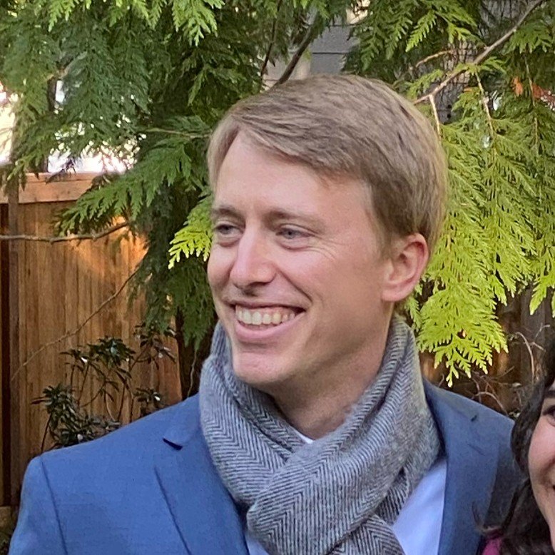 Smiling man with light brown hair wearing a blue blazer, gray scarf, and light shirt, outdoors with green leaves and a wooden fence in the background.