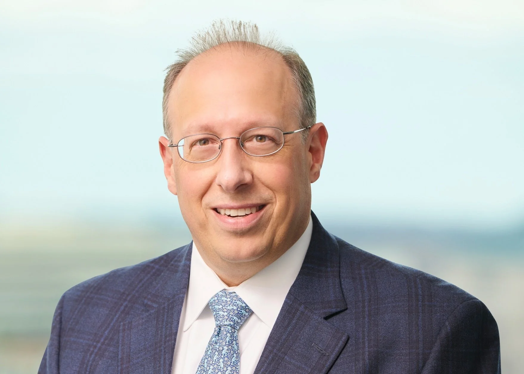 A smiling middle-aged man with glasses, wearing a dark checked suit, white shirt, and patterned tie, in front of a blurred outdoor background.