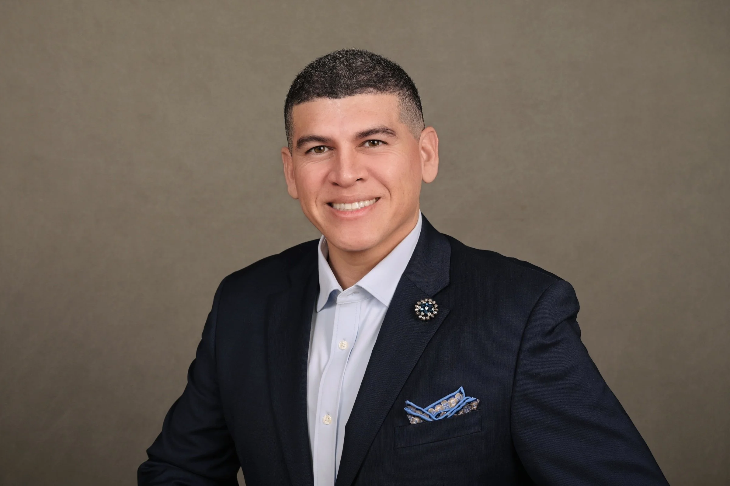 Portrait of a young man with short dark hair, wearing a dark suit, white shirt, and a decorative lapel pin, smiling in front of a plain background.