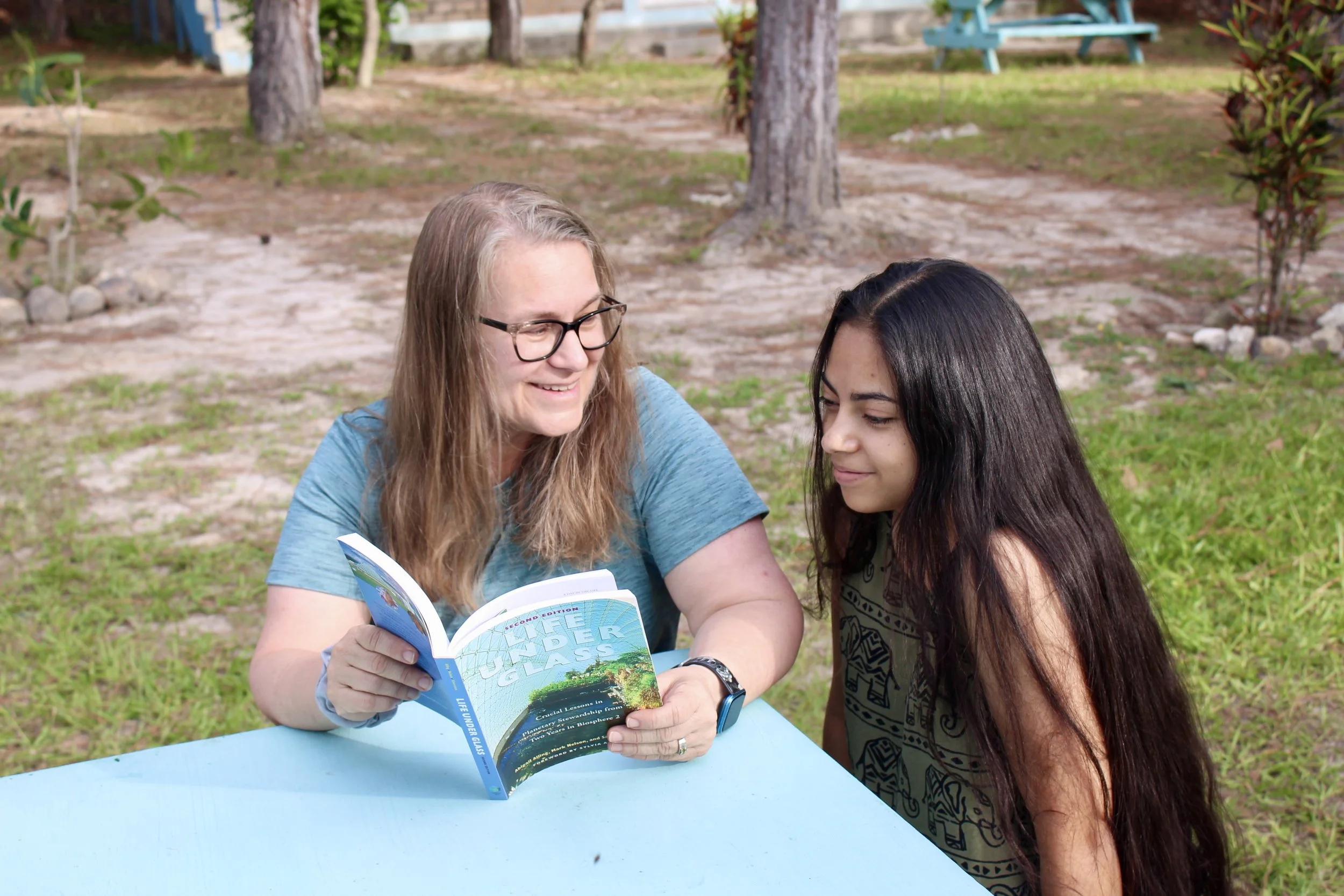 Two women, one with glasses and long wavy hair, the other with long dark straight hair, sit at a blue outdoor table in a park, reading a blue book together. Trees and a picnic table are in the background.