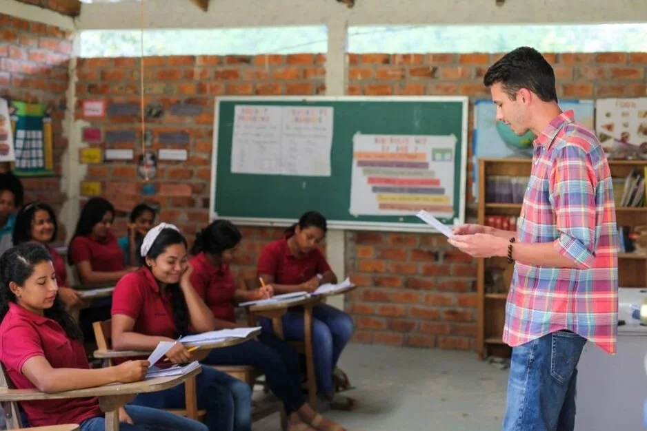 Teacher standing and reading from paper to a classroom of seated students, who are wearing red shirts and taking notes, in a classroom with brick walls and a blackboard.