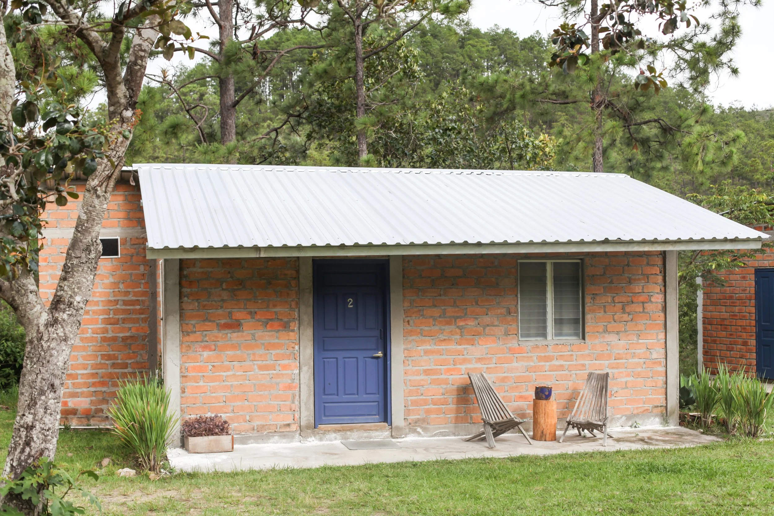 A brick house with a blue door and a dark window, front porch, surrounded by greenery and trees, with outdoor chairs and plants in front.