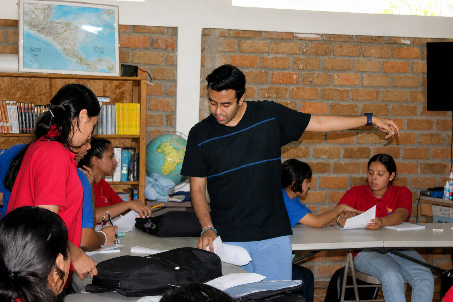 Teacher standing and pointing at students in a classroom with brick wall, bookshelves, a globe, and students reading or writing at desks.