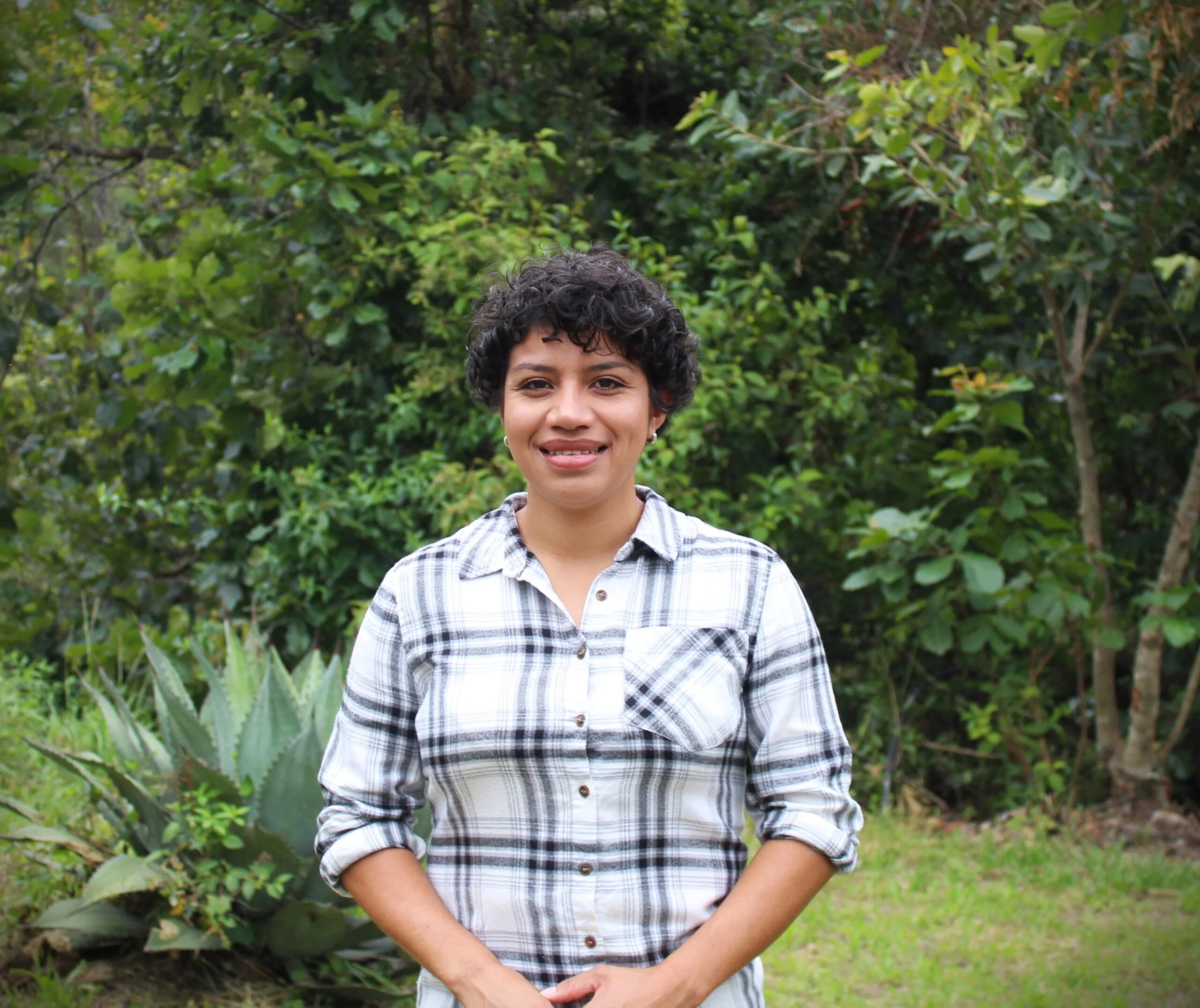 Young person with curly dark hair smiling outdoors in front of lush green foliage, wearing a plaid button-up shirt with rolled-up sleeves.