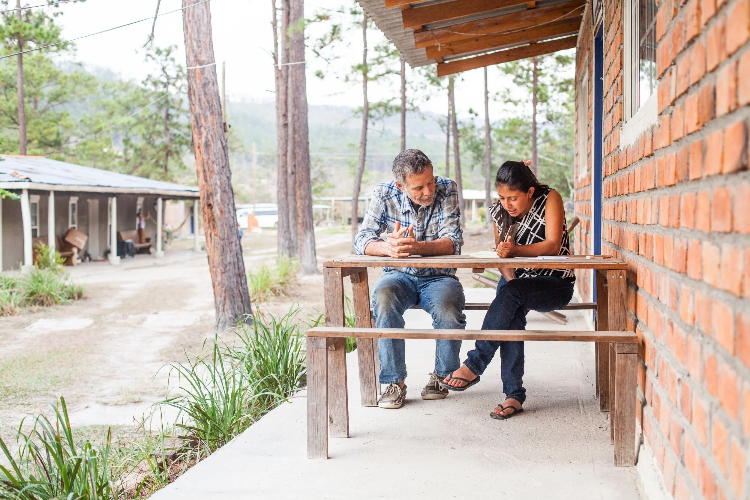 An older man and a young woman sit at a wooden table on a porch outside a brick house, looking at a phone together. The porch has a brick wall, and there are trees and other houses in the background.