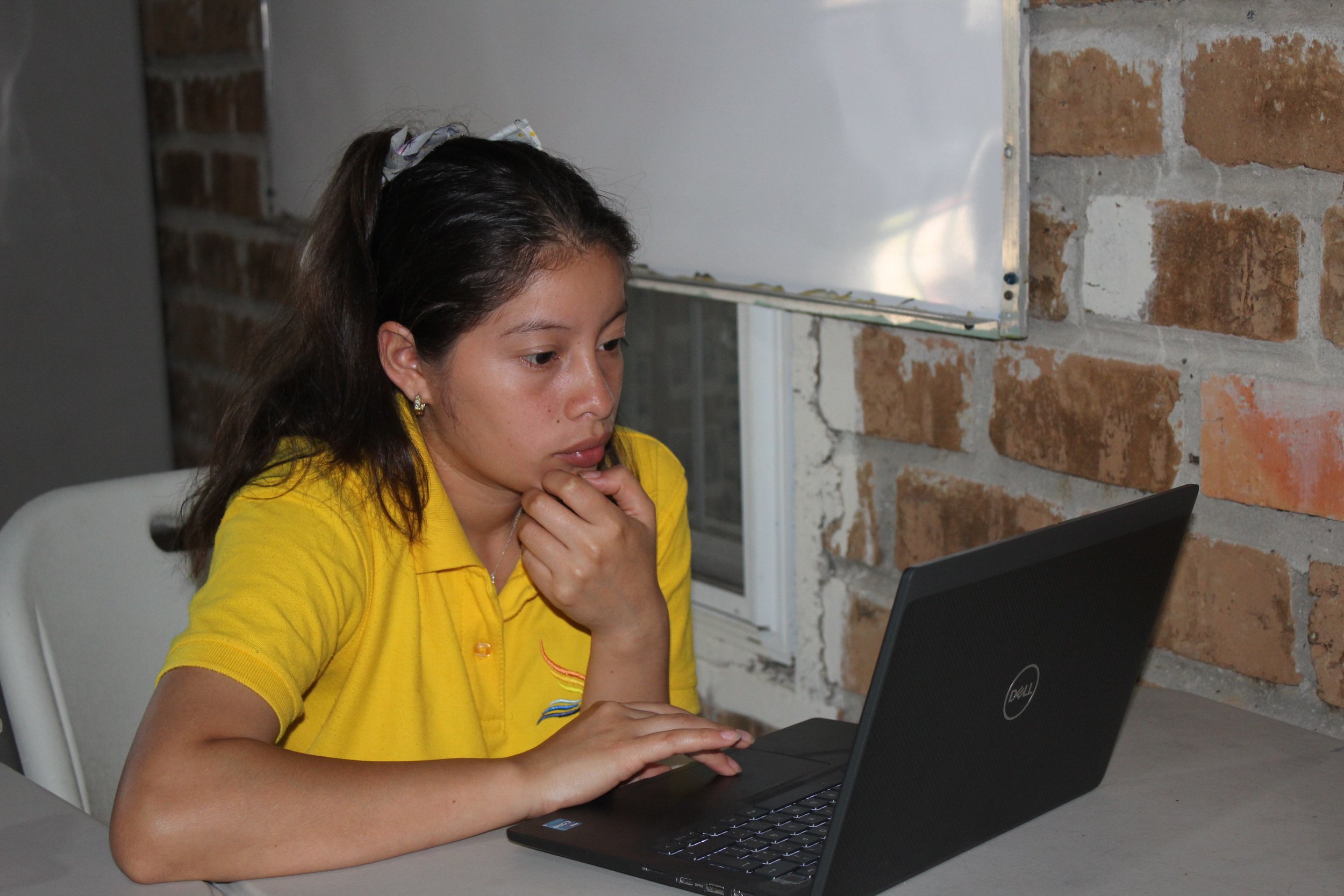 A young woman in a yellow shirt sitting at a gray table using a black laptop in front of a brick wall.