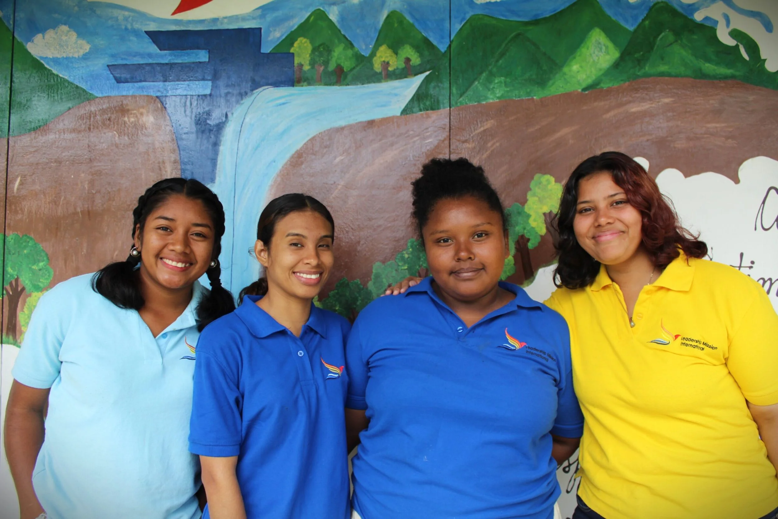 Four young women are standing in front of a colorful painted mural of mountains, trees, clouds, and a waterfall. They are smiling and wearing uniforms with logos, with the woman on the right in a yellow shirt and the others in blue shirts.
