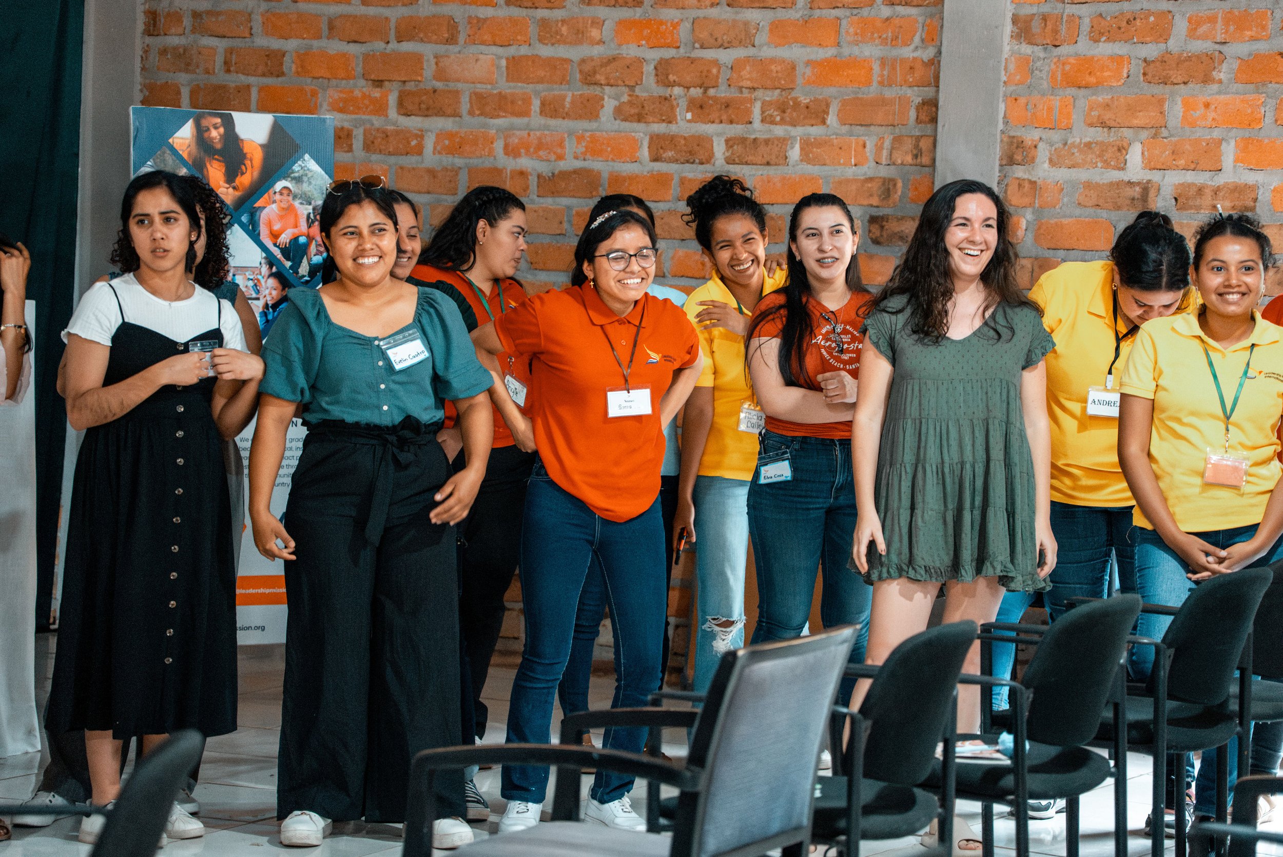 A group of diverse young women standing together at an indoor event, smiling and laughing, with a brick wall in the background.