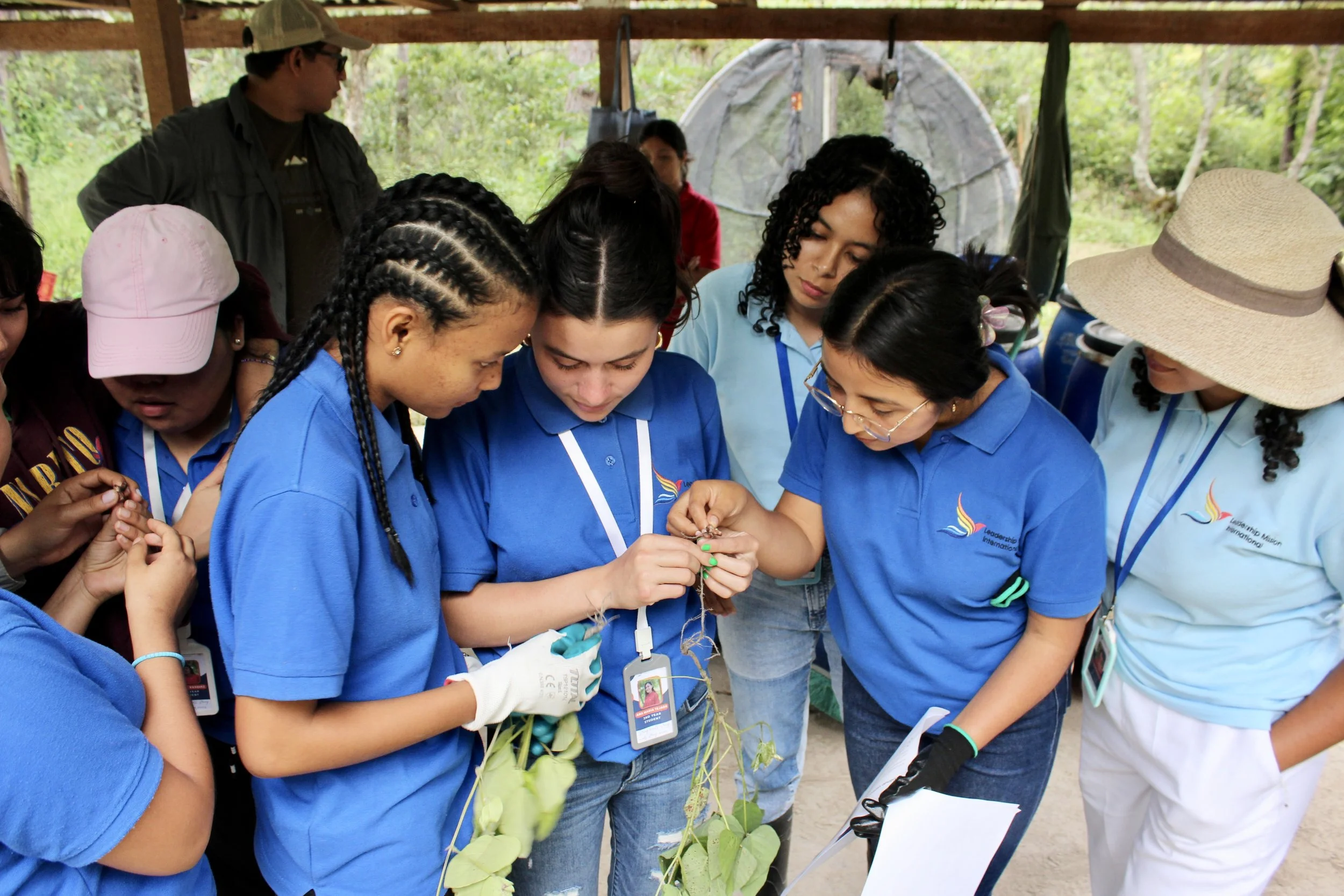 A group of young women wearing blue shirts with a logo, examining plants together outdoors, with some wearing gloves. A man in a cap and sunglasses is observing beside them. The setting appears to be an outdoor educational or conservation activity surrounded by greenery.