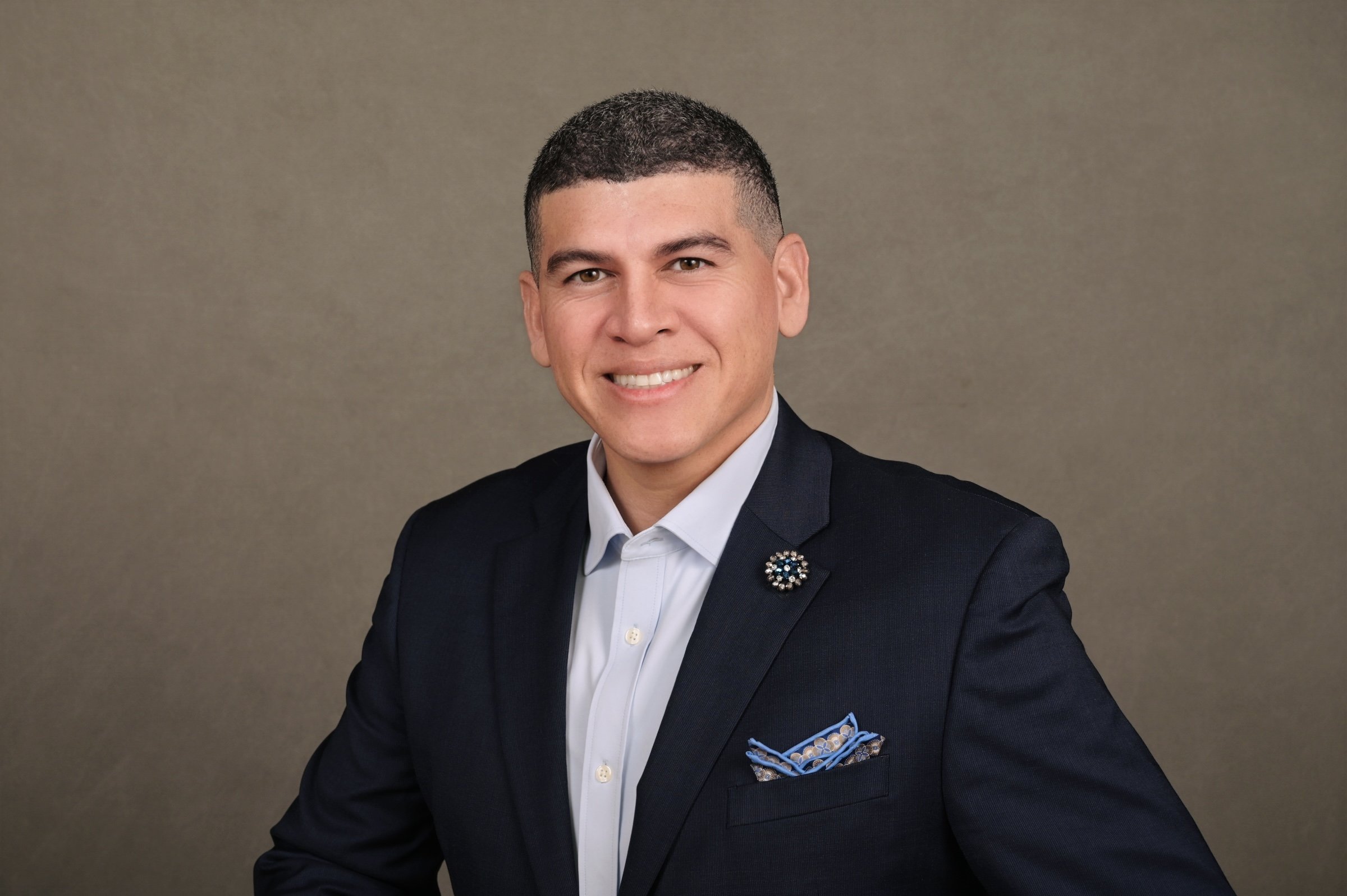 Smiling man wearing a dark suit, white shirt, and a decorative lapel pin, posed against a neutral background.