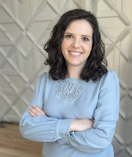 Woman with dark wavy hair smiling and crossing her arms, wearing a light blue blouse with lace detail, standing in front of a beige geometric wall.
