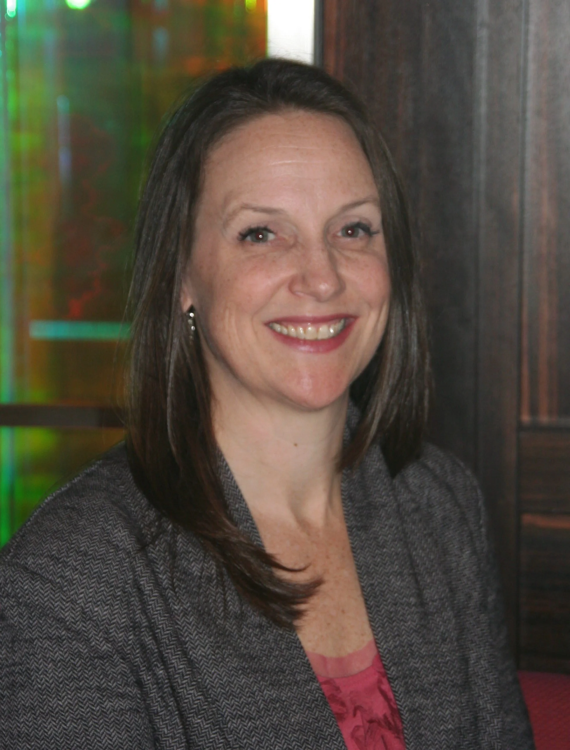 A woman with shoulder-length dark hair, wearing a gray blazer and a pink blouse, smiling in front of a colorful, blurred background.