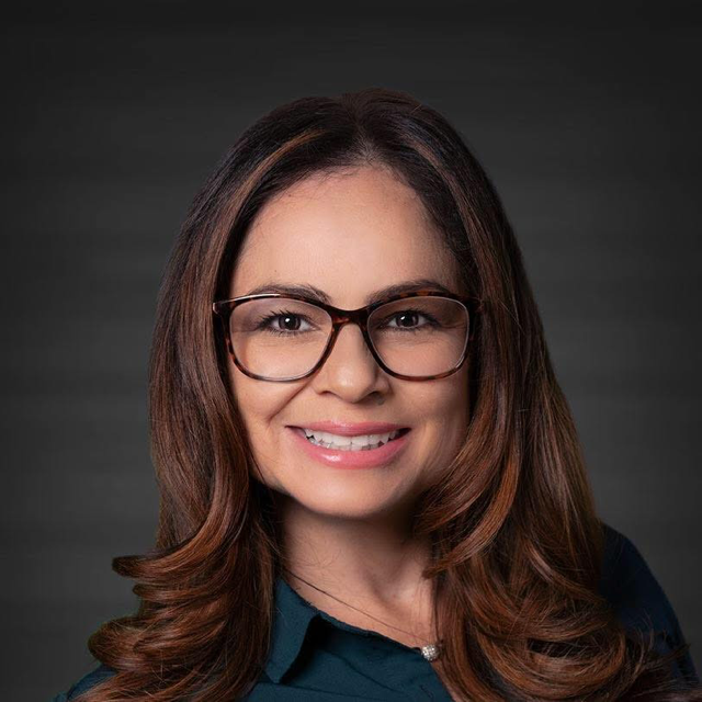 A smiling woman with long brown hair, wearing glasses and a dark blue top, against a dark gray background.