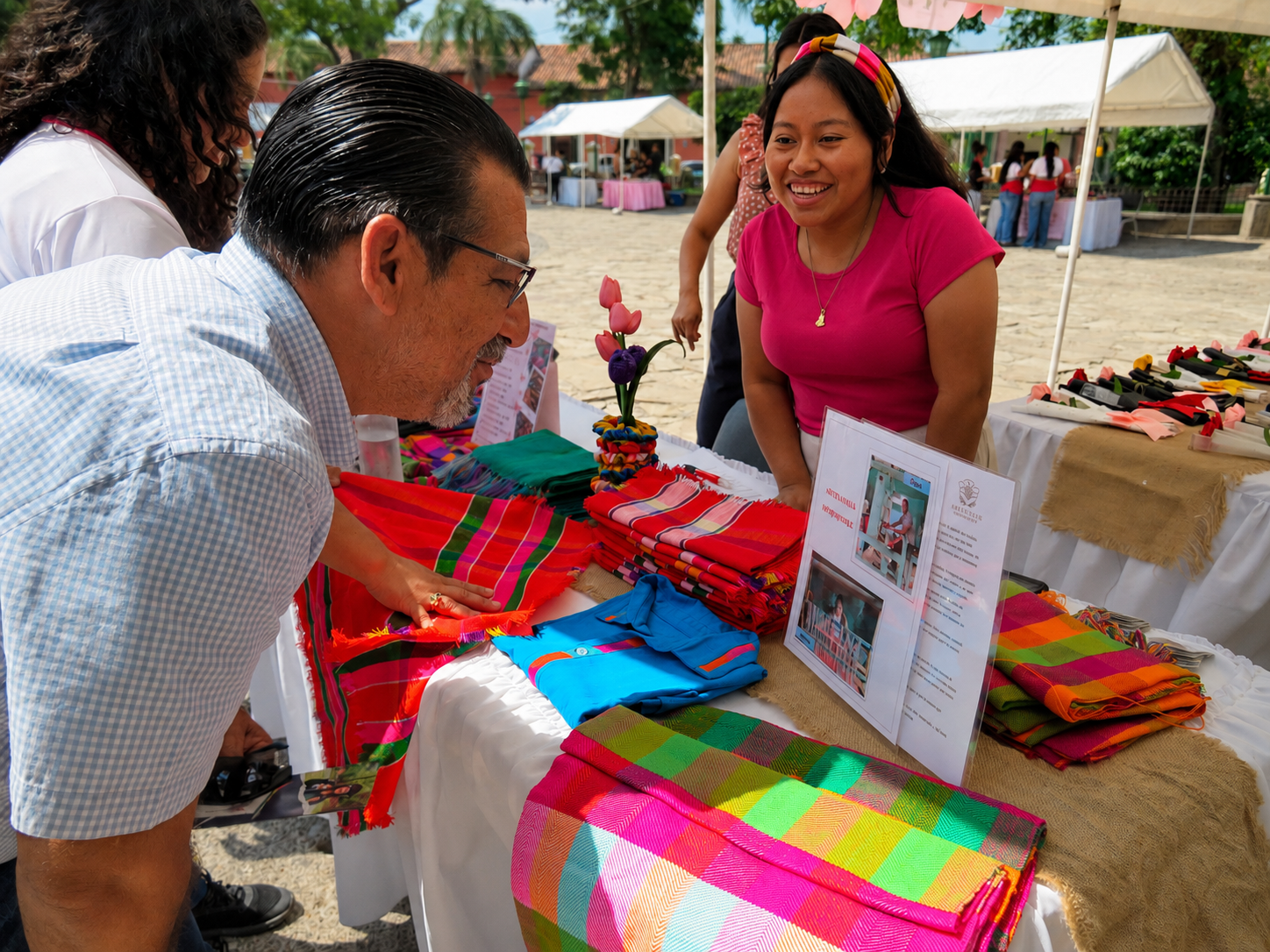 A woman in a pink shirt smiling at a man at a colorful outdoor market stall. Various brightly colored scarves and textiles are displayed on the table, with a decorative flower arrangement and informational signs visible. Other market stalls and people in the background.