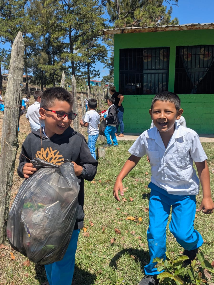 Two boys smiling and laughing outdoors near a green building, one holding a trash bag, surrounded by other children and trees.