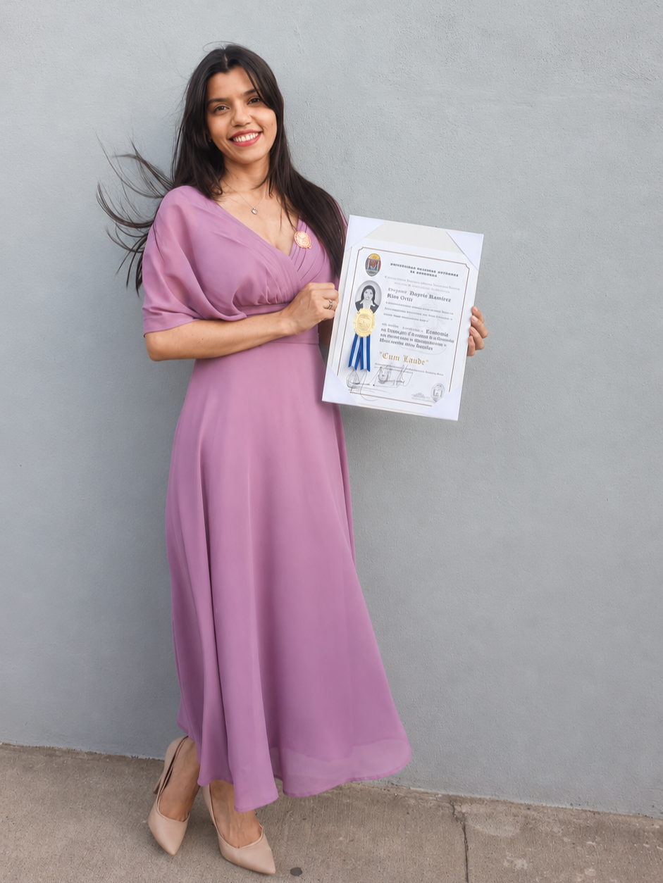 A young woman in a purple dress holding a certificate of achievement and smiling at the camera, standing against a light gray wall.