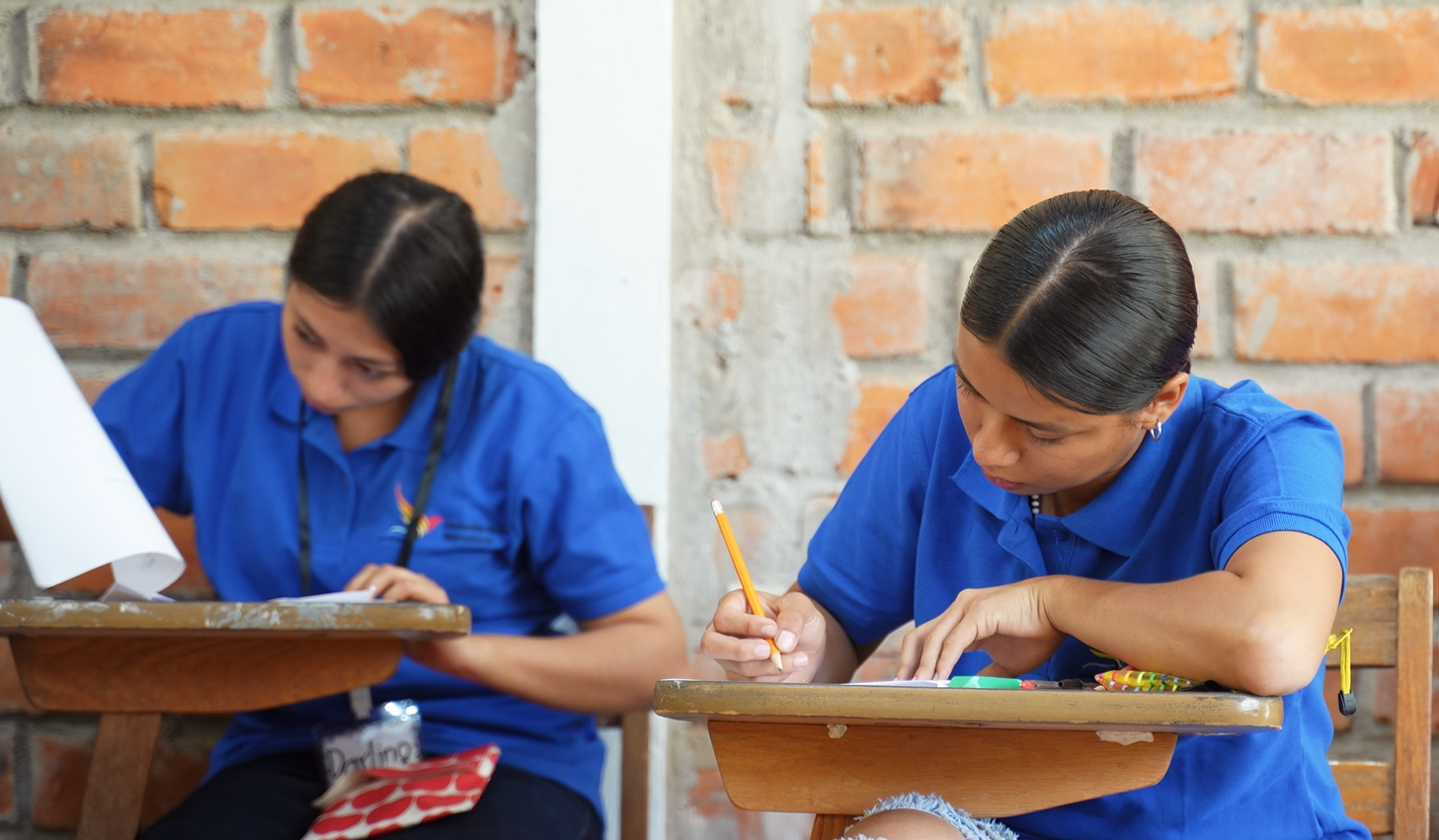 Two girls in blue shirts sitting at desks writing on paper with a brick wall background.