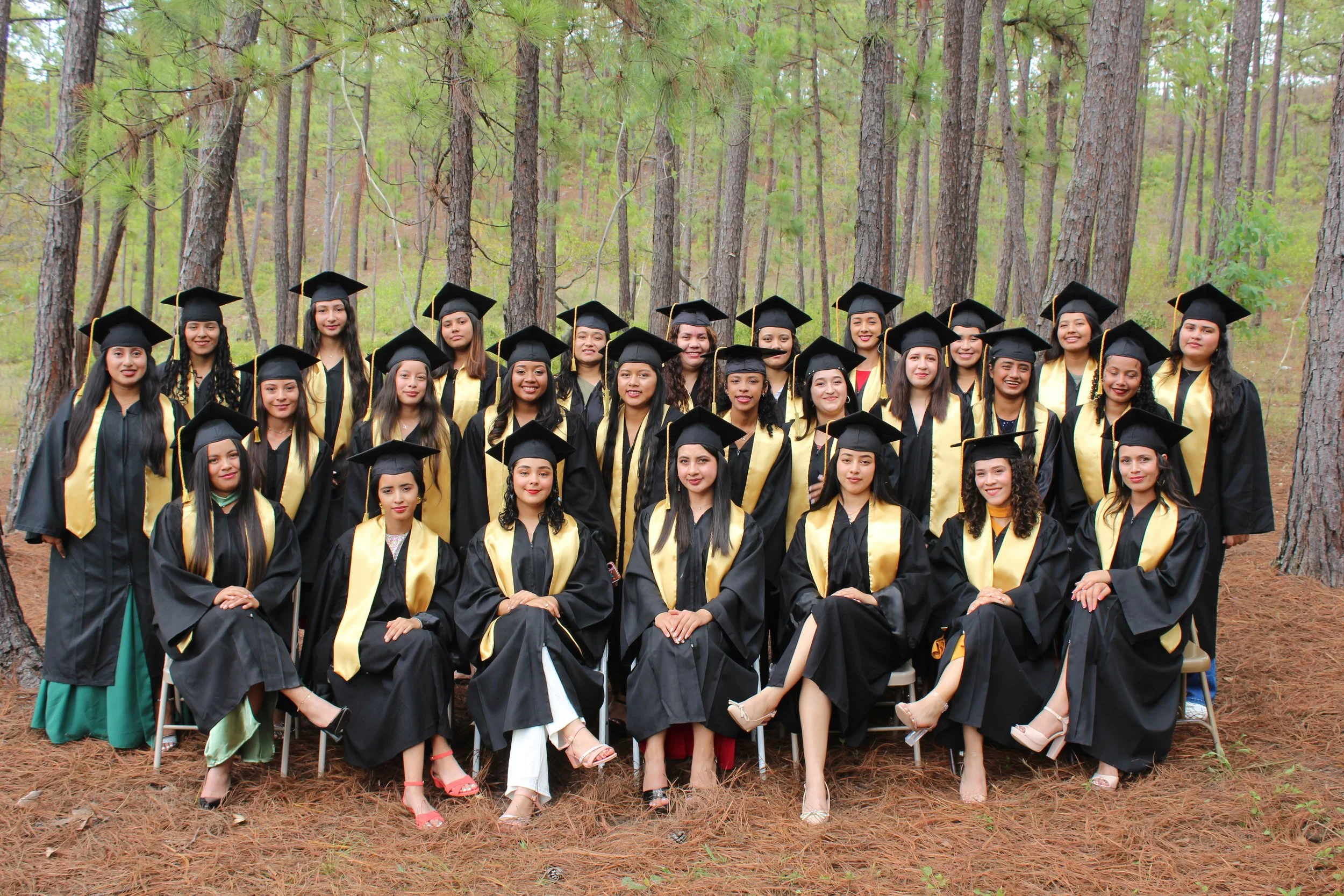 Group of young women dressed in graduation caps and gowns posing outdoors in a forested area.
