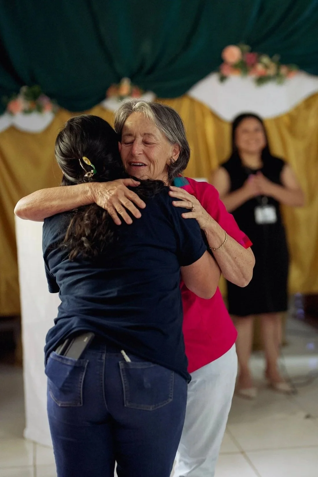 Two women hugging warmly in a room with a decorated background. One woman has gray hair and is wearing a pink top. The other woman has long dark hair with a hair clip, wearing a dark top. A third woman in a black dress is in the background, smiling and clapping.