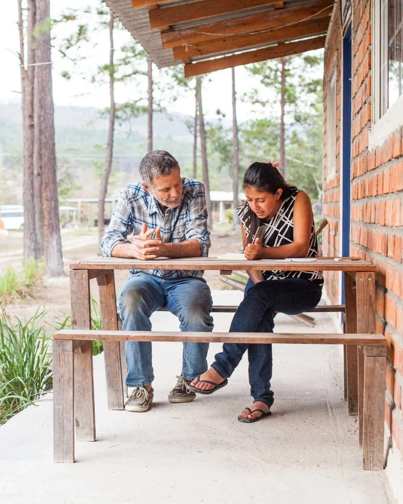 A young woman and an older man sitting at a wooden outdoor table on a porch, with the woman showing something on her phone to the man, both looking at it intently.