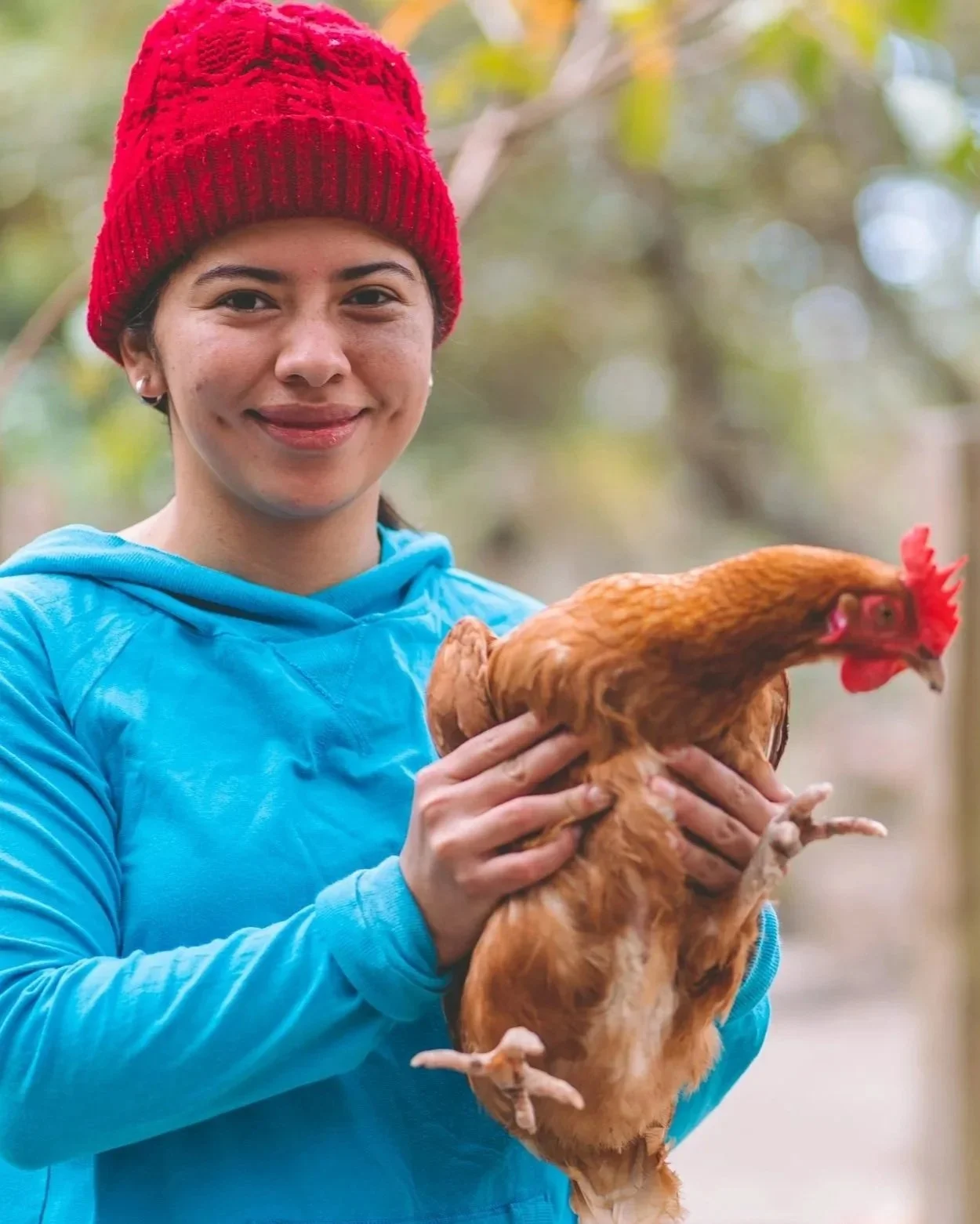 A woman wearing a red knit hat and a blue jacket holding a brown chicken in an outdoor setting.