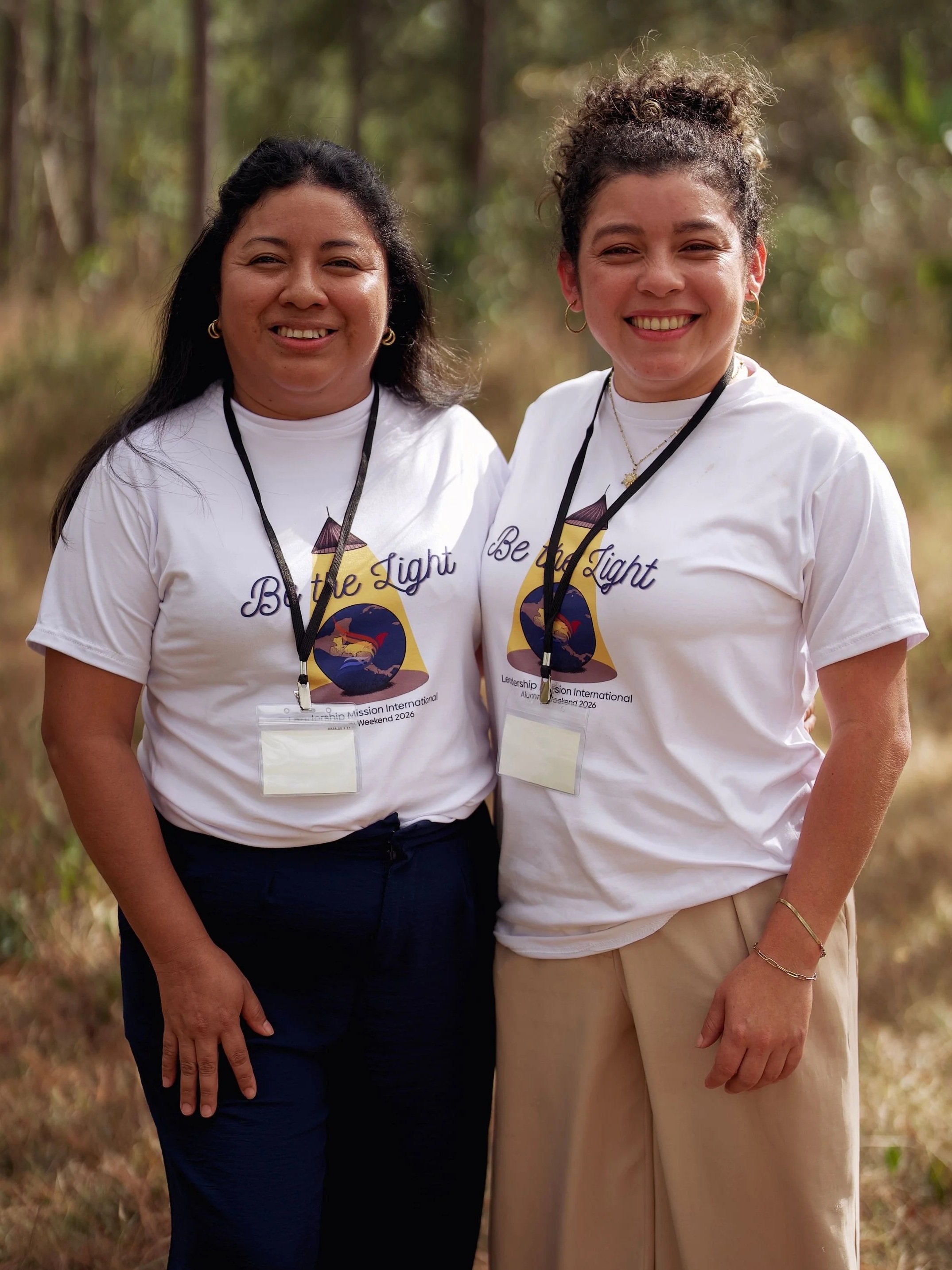 Two women standing outdoors in a natural setting, smiling at the camera, wearing white t-shirts with a logo and text that reads 'Be the Light,' along with conference or event badges around their necks.