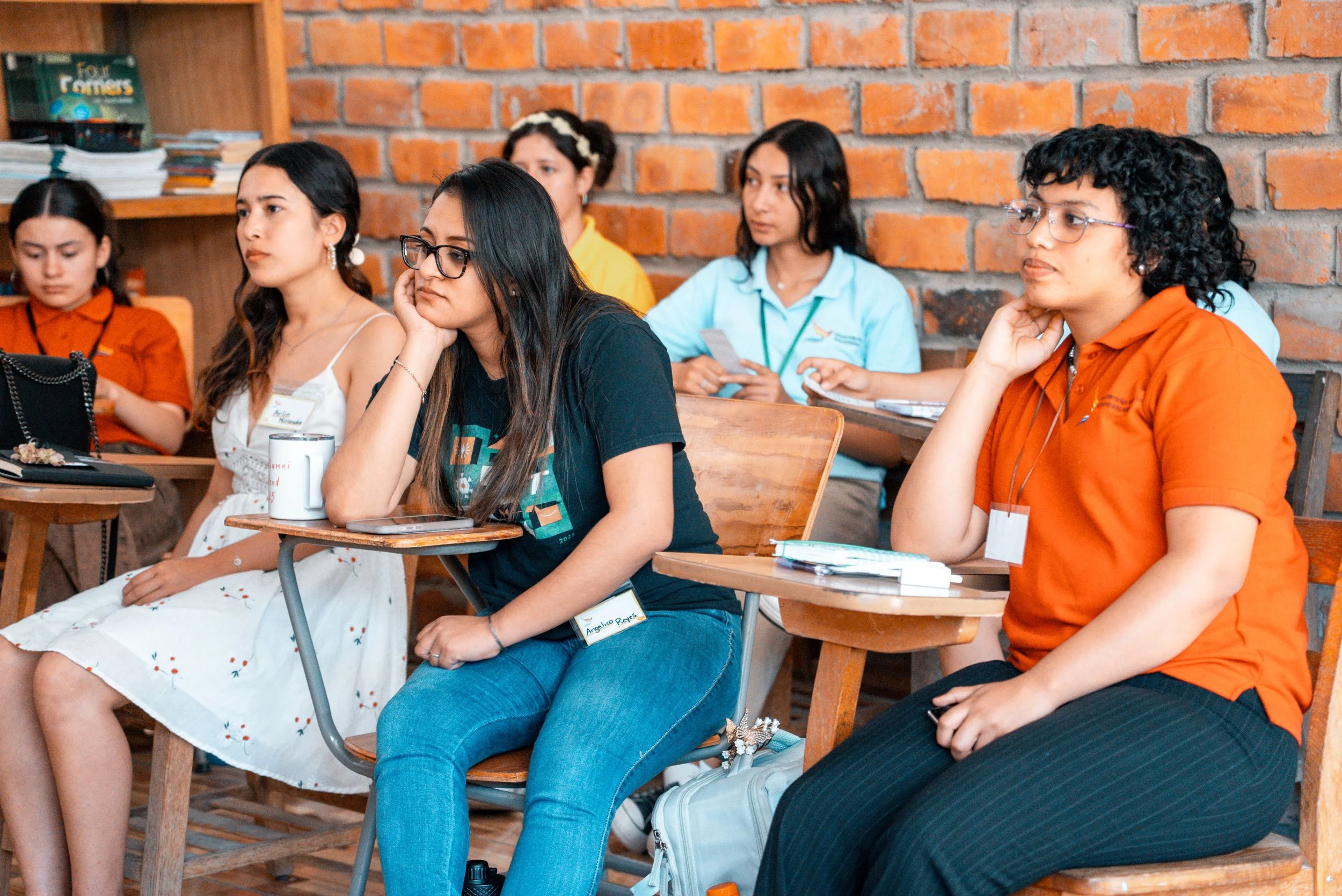 Group of young women sitting at desks in a classroom or workshop, listening attentively, with a brick wall background.