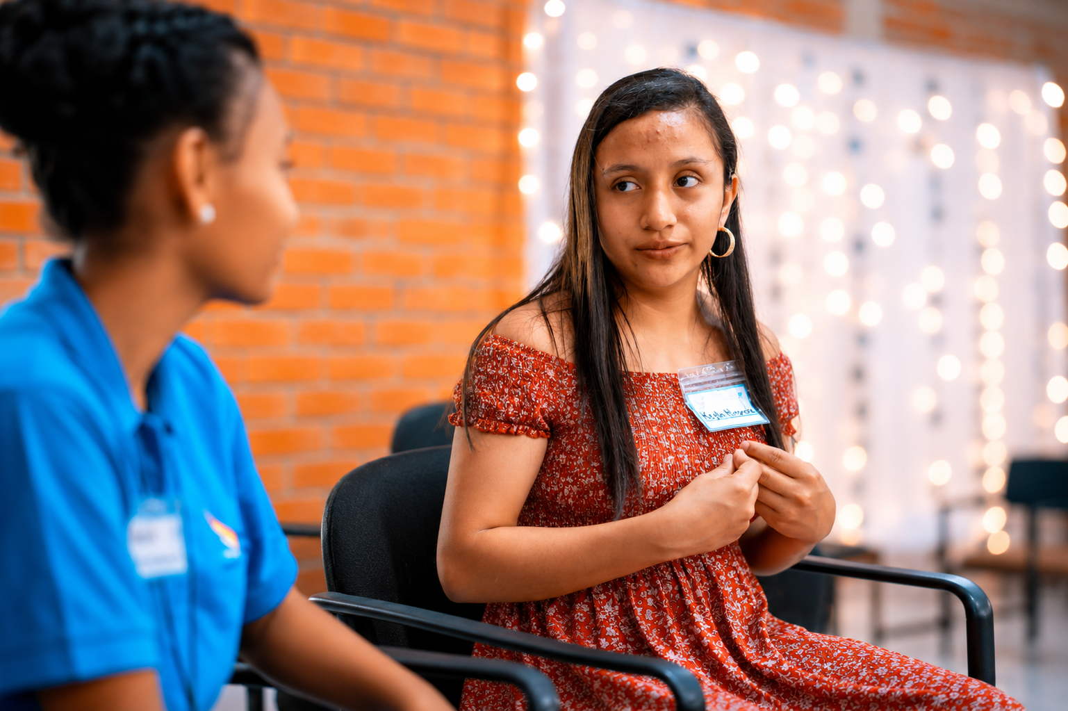 A young girl with long dark hair wearing a red patterned dress and hoop earrings sits in a chair, engaging in a conversation with a woman dressed in a blue shirt. The setting appears to be indoors, with a brick wall and string lights in the background.