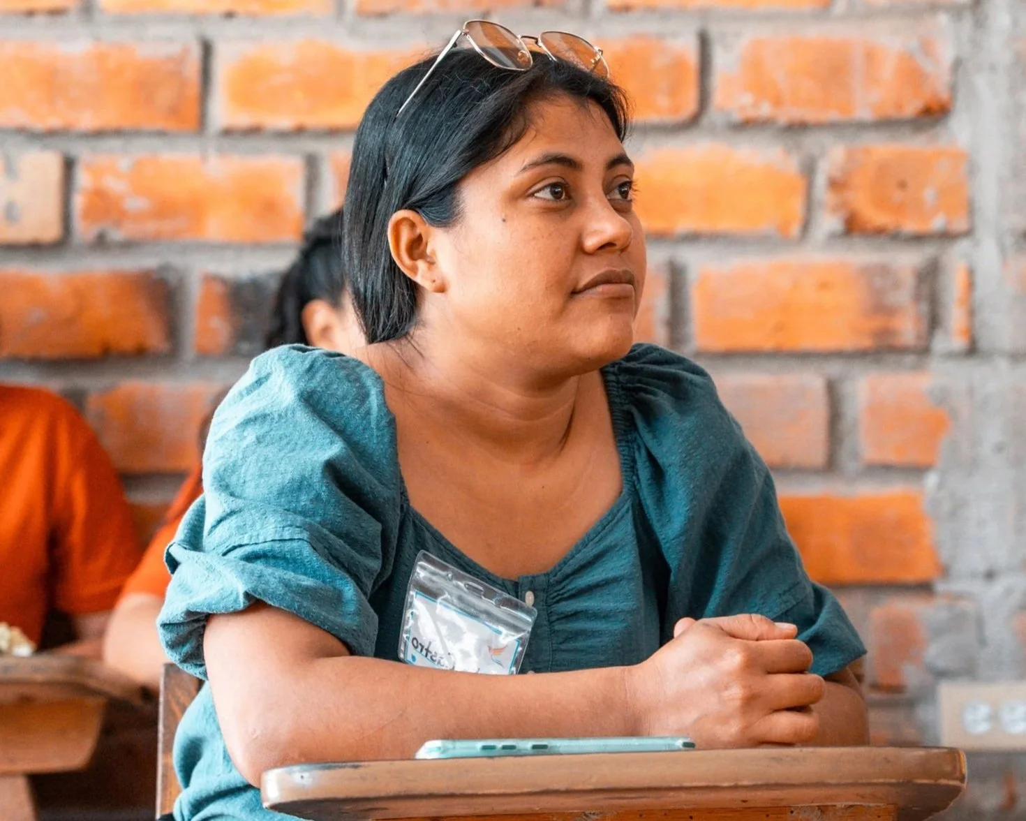 A woman with short black hair and sunglasses on her head sits at a wooden table with a brick wall in the background.