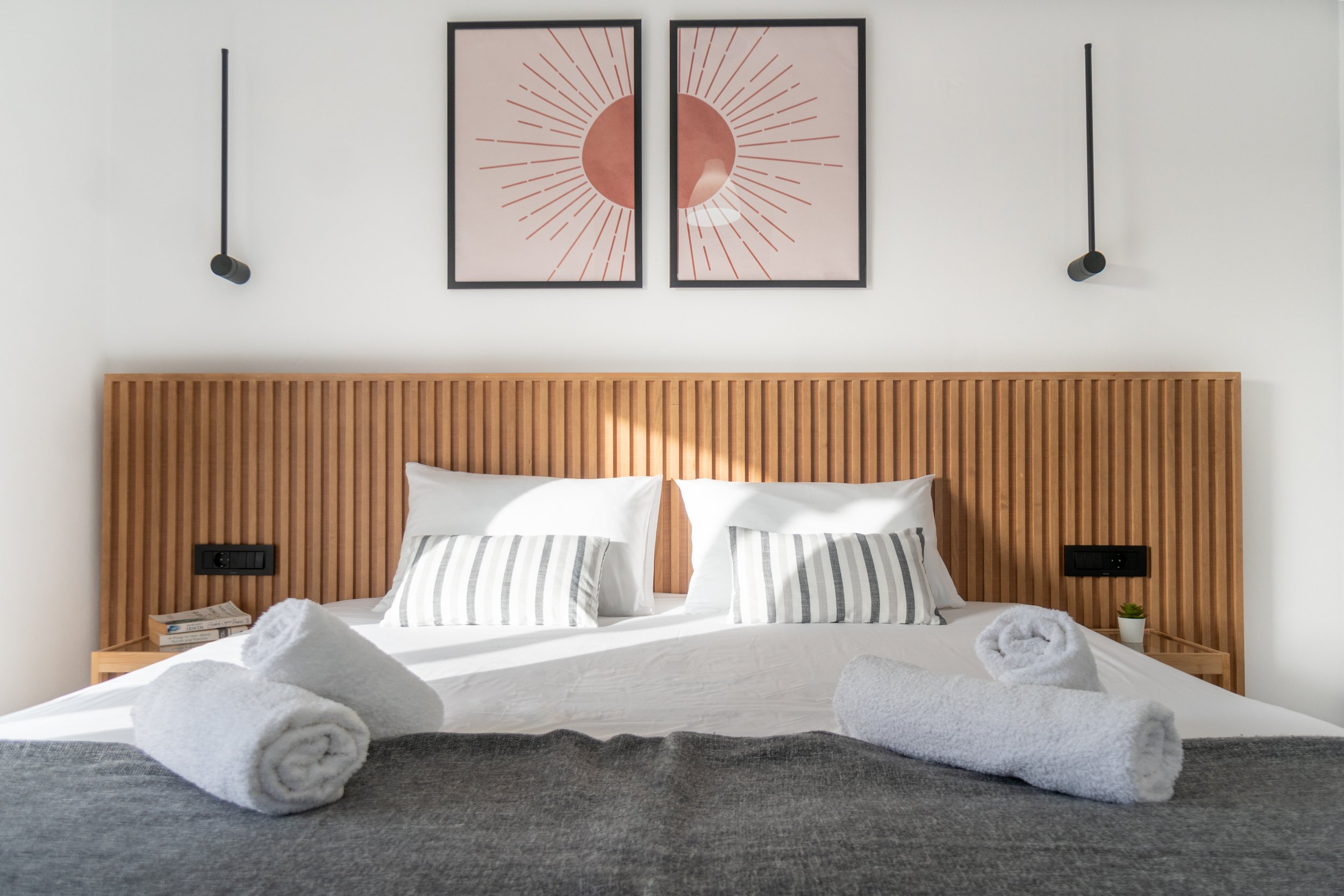 A neatly made double bed with striped pillows, with two rolled white towels placed on the bed and a gray blanket at the foot. The bed has a wooden slatted headboard. Above the bed are two modern black-framed abstract art prints featuring a large circle with radiating lines, hung on a white wall. There are two black wall-mounted reading lamps, one on each side of the art, with one lamp turned off and the other reflecting light.