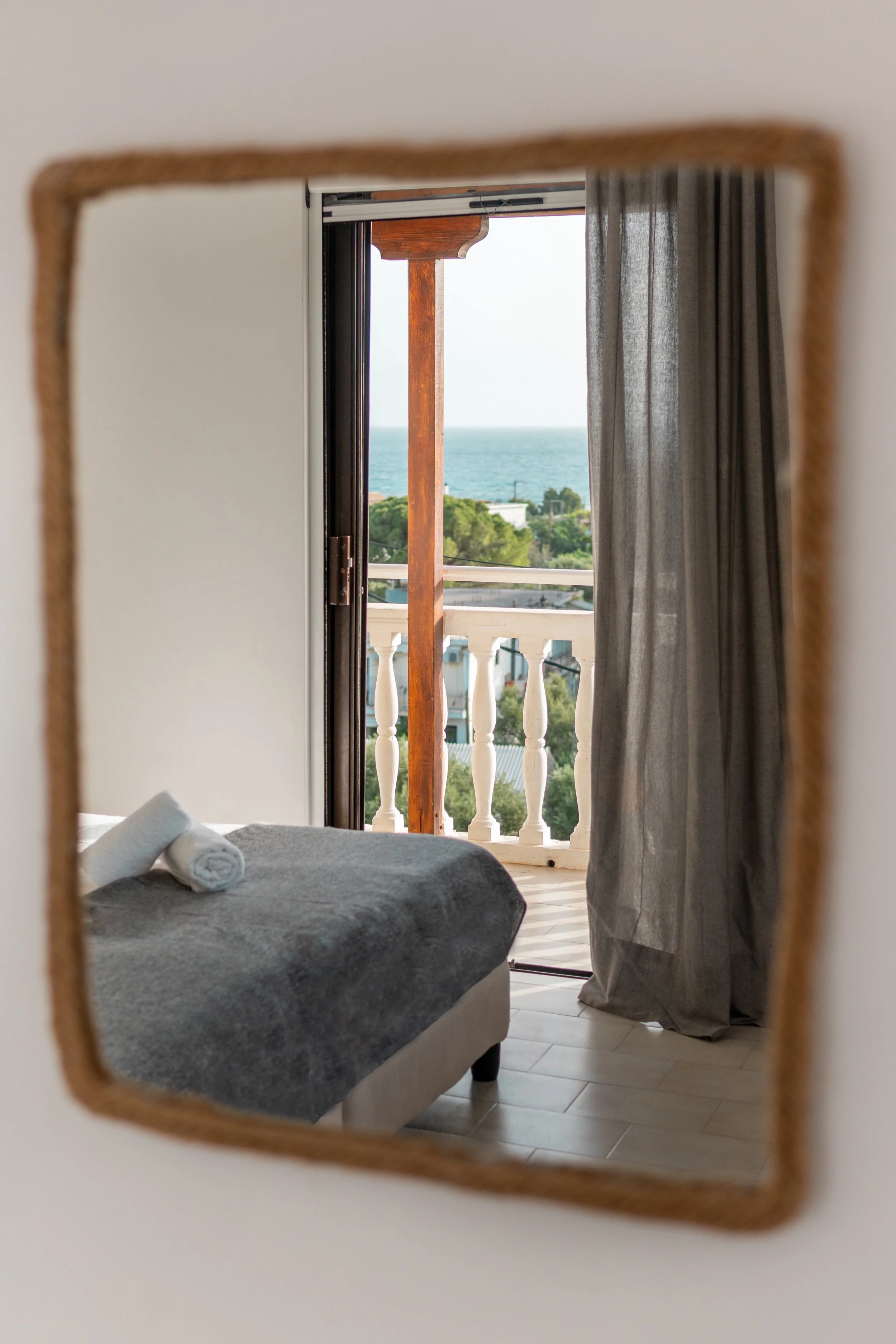 View through a mirror of a bedroom with a bed, two rolled towels, a door opening to a balcony, with curtains, overlooking trees, buildings, and a body of water in the distance.
