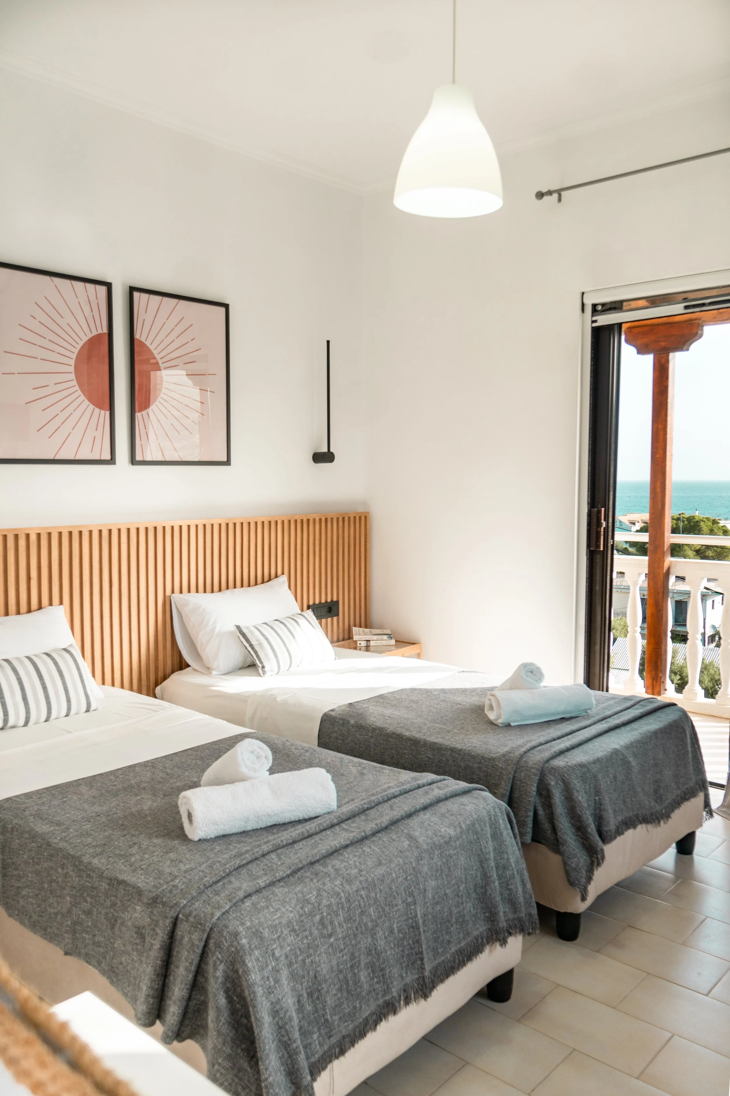 Two twin beds with white linens, striped pillows, and rolled towels on grey blankets in a bright room with a balcony view of the ocean.
