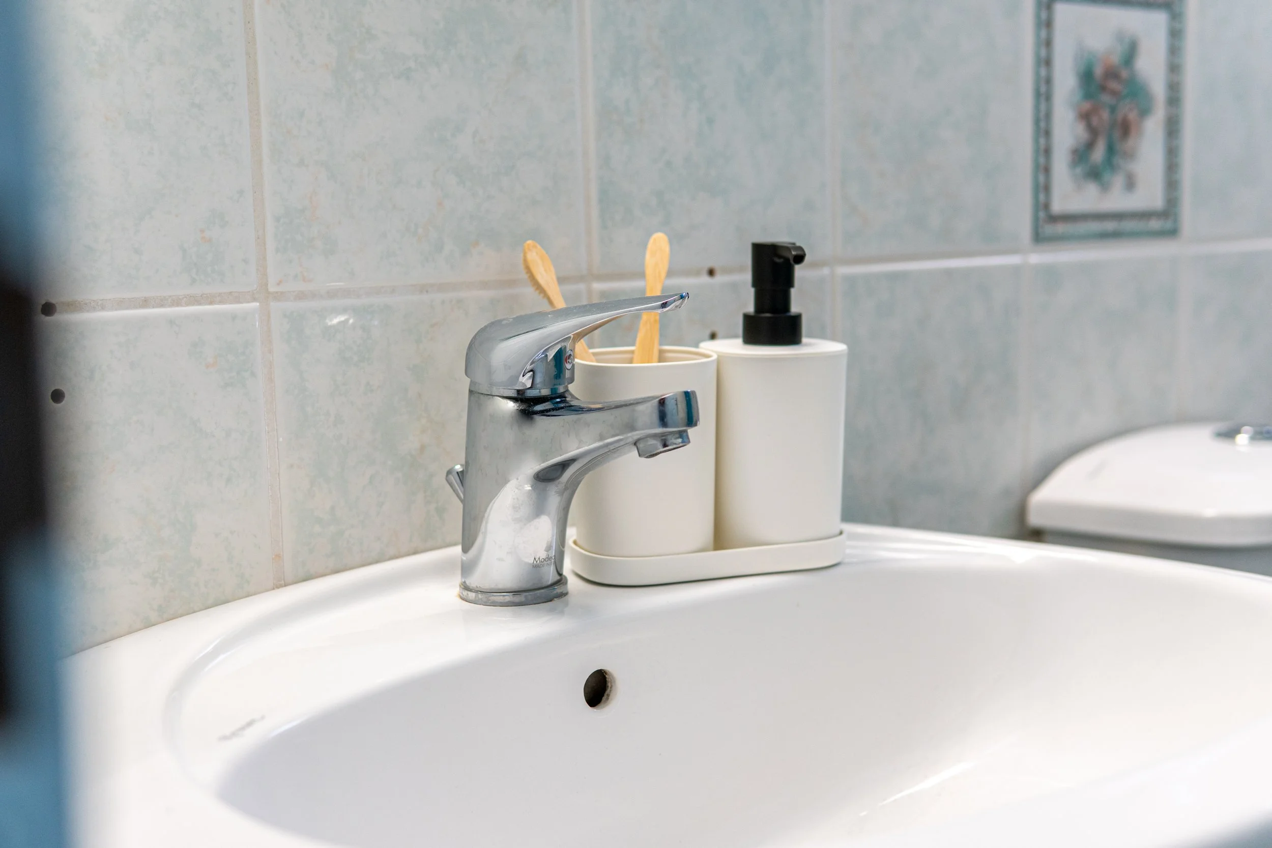 Close-up of a bathroom sink with a silver faucet, a soap dispenser, and two toothbrushes in a holder, against a tiled wall.