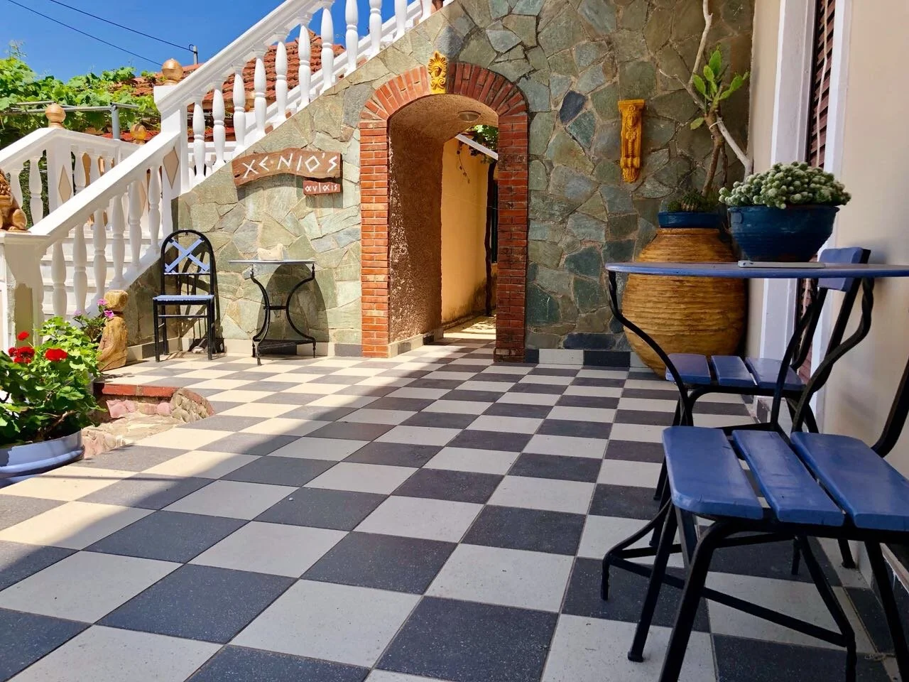 A patio with black and white checkered tile flooring, surrounded by a stone and brick wall. There are black chairs and tables, potted plants, and a staircase with white balusters leading up on the left.