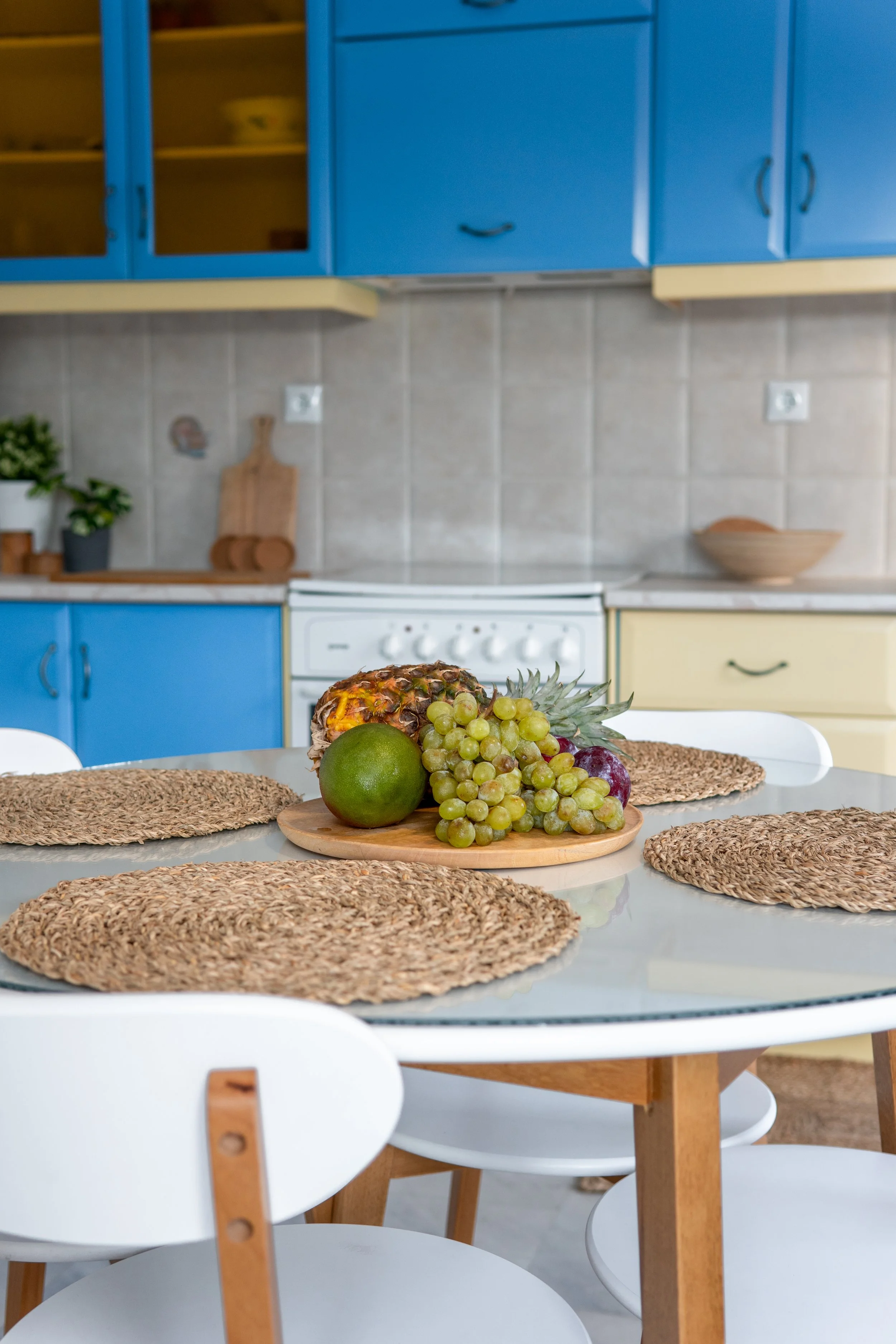 Kitchen with blue and yellow cabinets, a white stove, countertop with a wooden cutting board, green and purple grapes, pineapple on a round wooden tray, and woven placemats on a white dining table with white chairs.