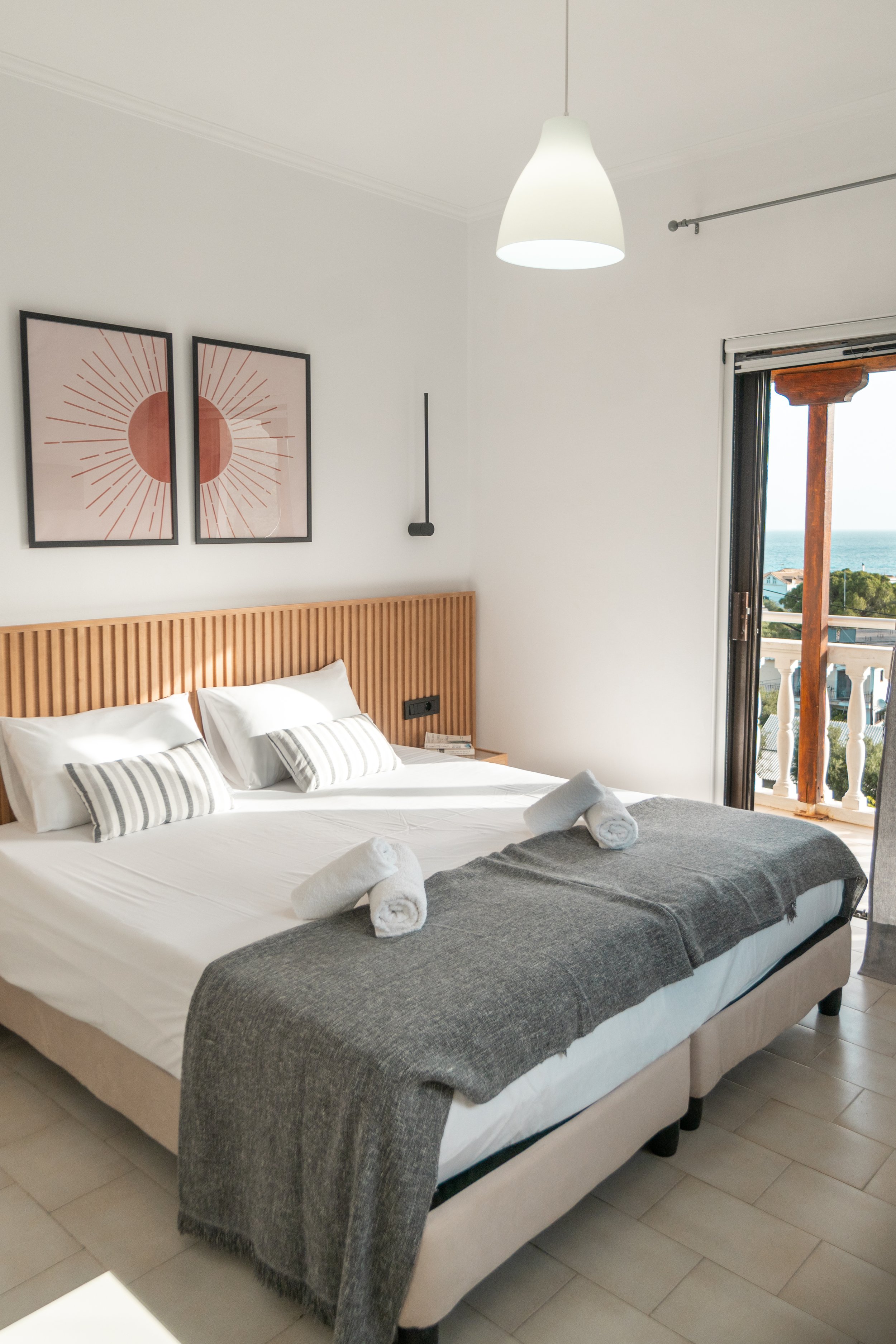A neatly made hotel bed with white sheets, striped pillows, and rolled towels on a dark gray blanket, with a wooden headboard, artwork on the wall, and an open balcony door revealing a seaside view.