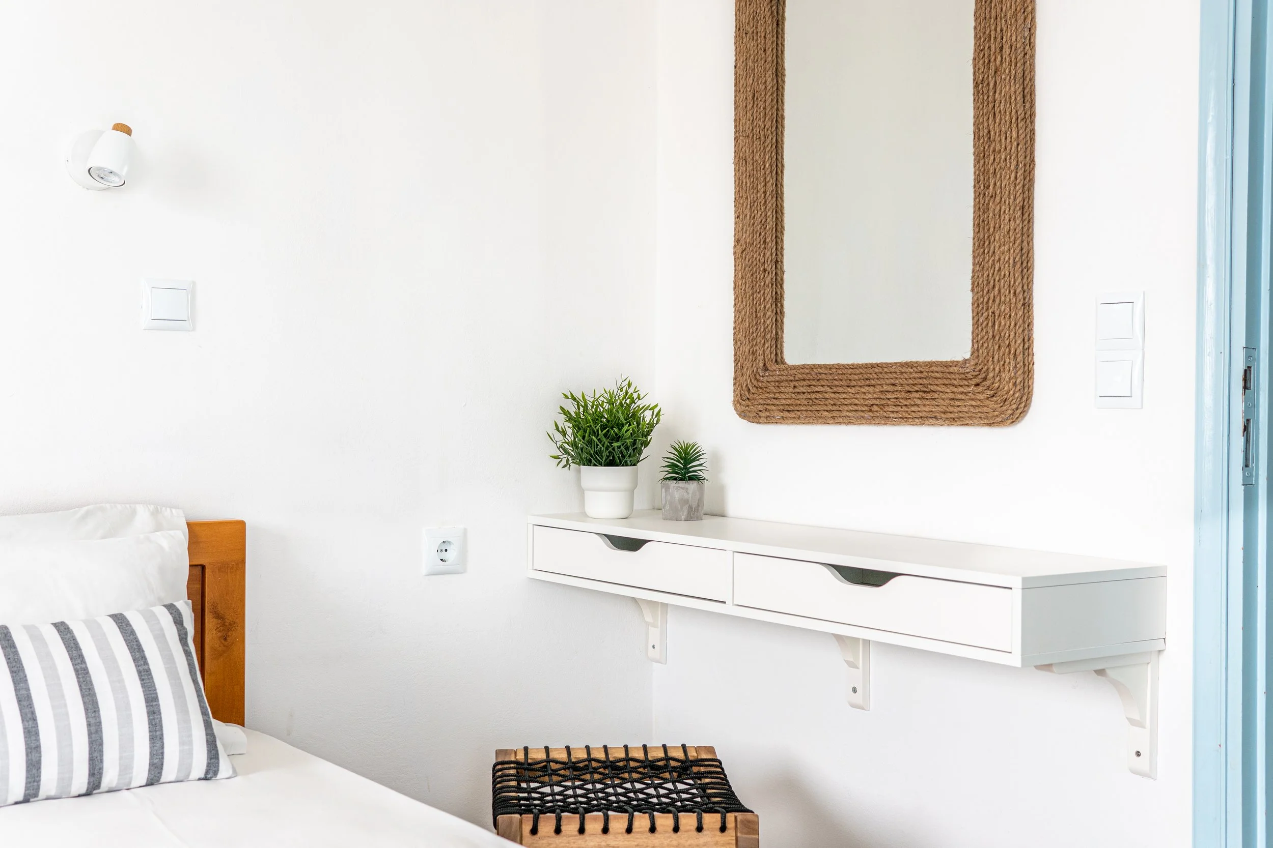 A bedroom corner featuring a white bed with a gray striped pillow, a white wall-mounted shelf with two green potted plants, a woven mirror frame, a small black woven stool or wicker side table, and a wall sconce light with a wooden accent.