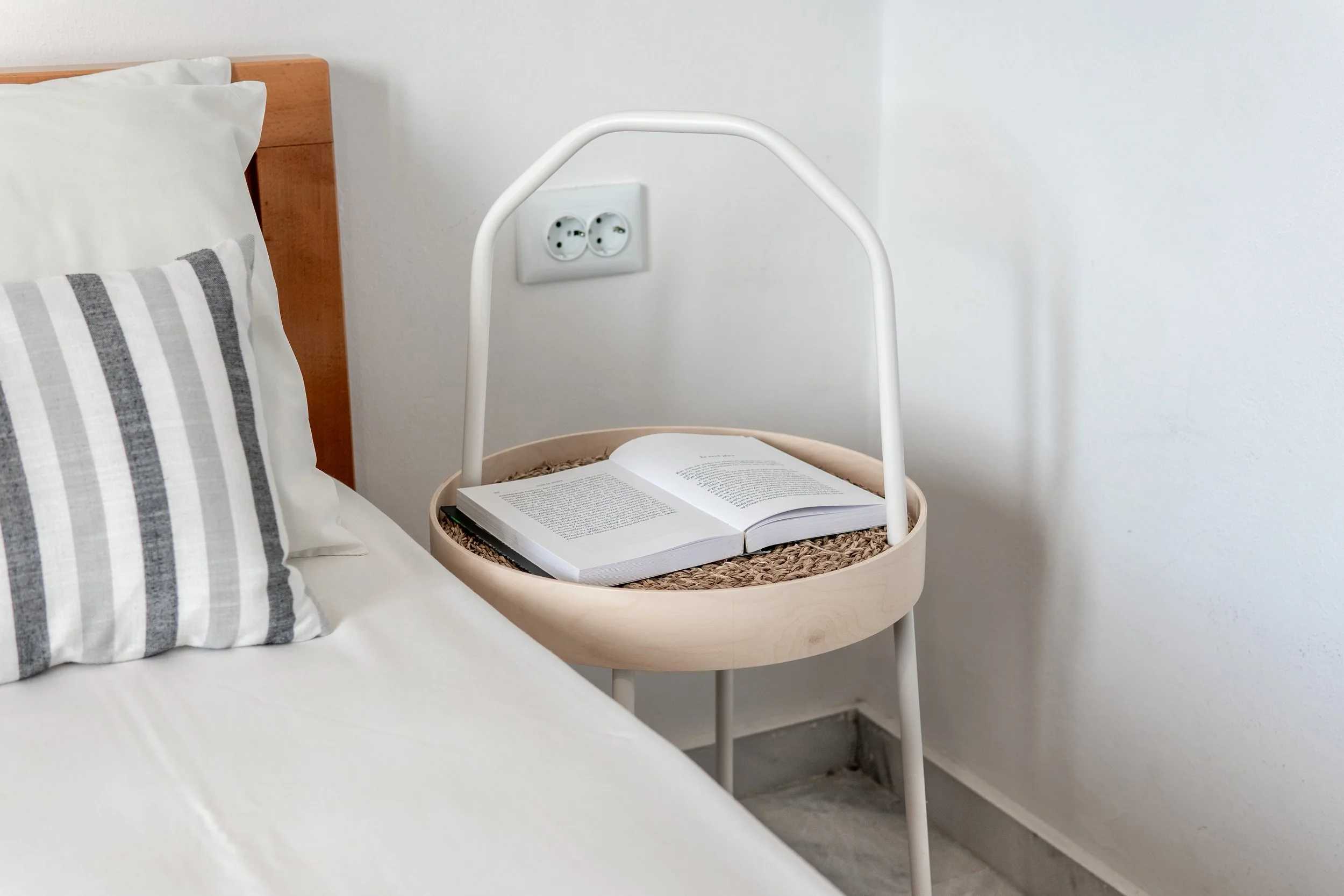 A bedroom with a white bed, striped pillow, and a beige bedside table with an open book and a white headboard. There is a white wall with electrical outlets.