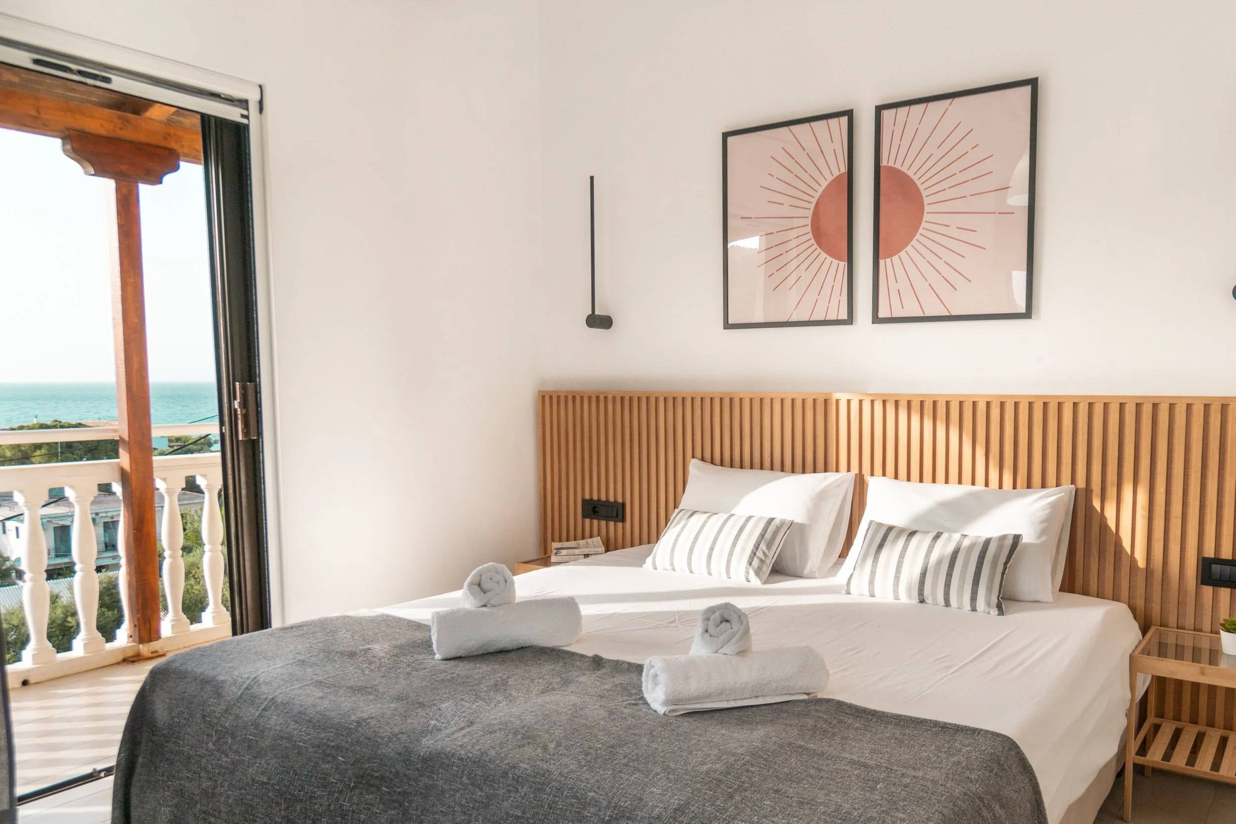 A bedroom with a bed, two pillows, towels, and a view of the ocean through an open balcony door. Two framed geometric art pieces hang above the bed, and a wooden headboard and nightstand are visible.