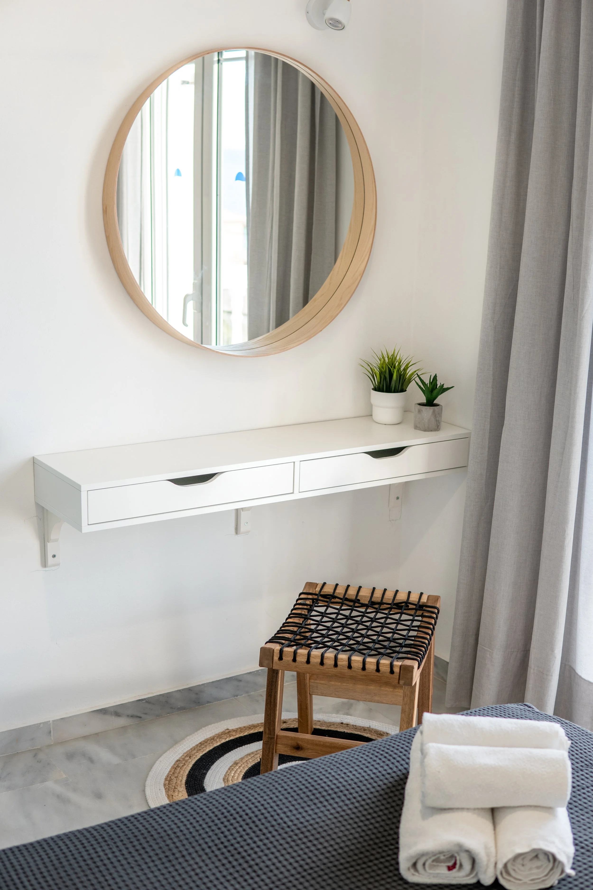 A bedroom scene with a mirror on a white wall, a small white shelf, and two green potted plants. Curtains are visible near the mirror, and a stool with a wooden frame and black woven top is on a circular rug. There are neatly rolled towels on a bed.