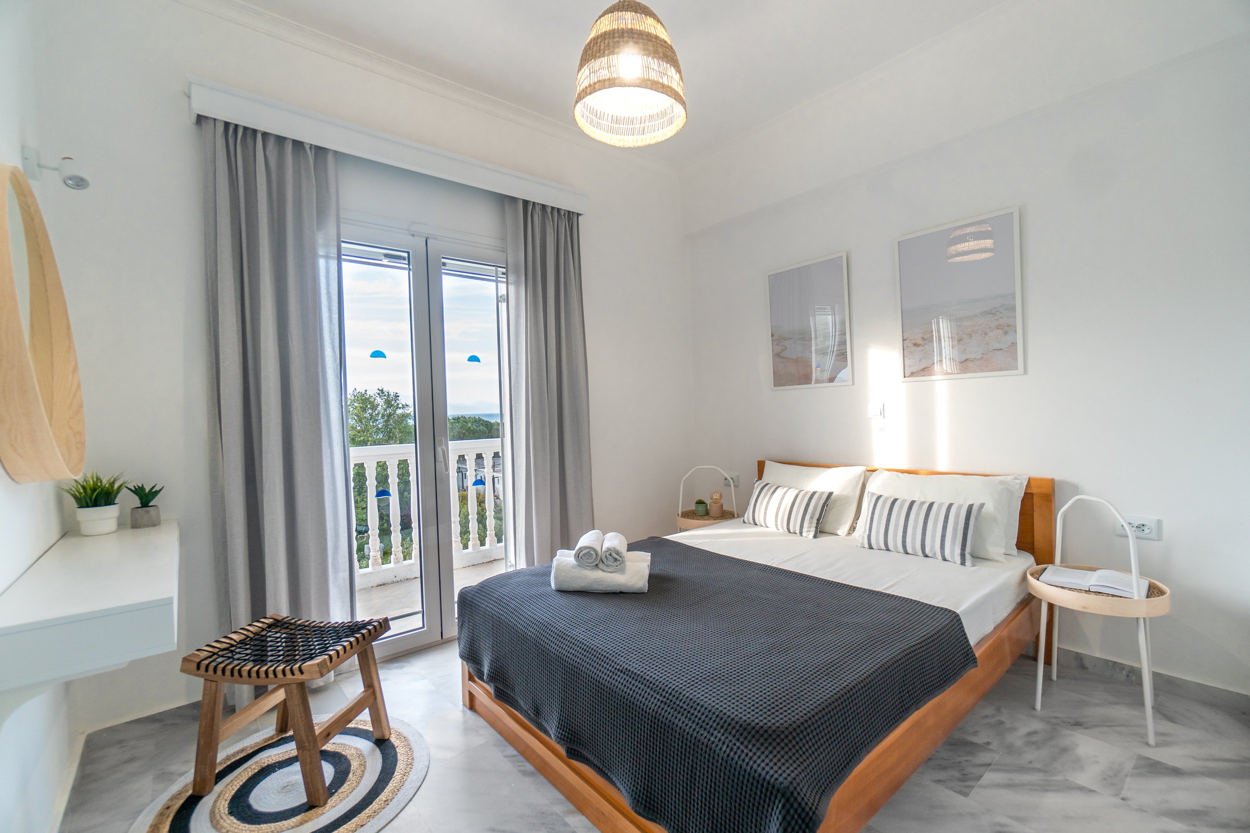 A bedroom with a large window and balcony door, gray curtains, framed beach pictures on the wall, a bed with striped pillows, a small round side table, and a woven stool at the foot of the bed.