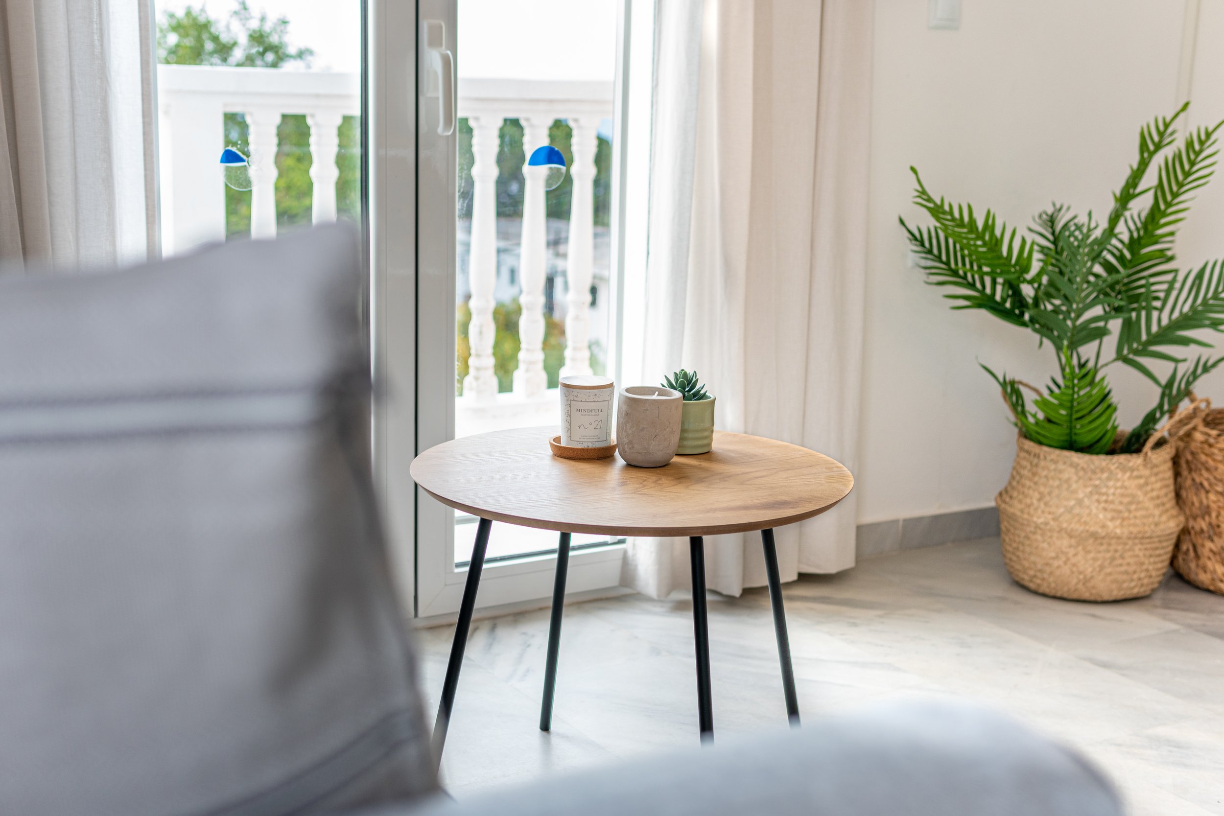 Interior scene with a wooden round table near a glass door. On the table are a white candle, a small succulent, and a taller beige candle holder. To the right, there is a green fern plant in a wicker basket. A white curtain and a balcony with white balustrades are visible through the glass door, with part of a white wall and green foliage outside.