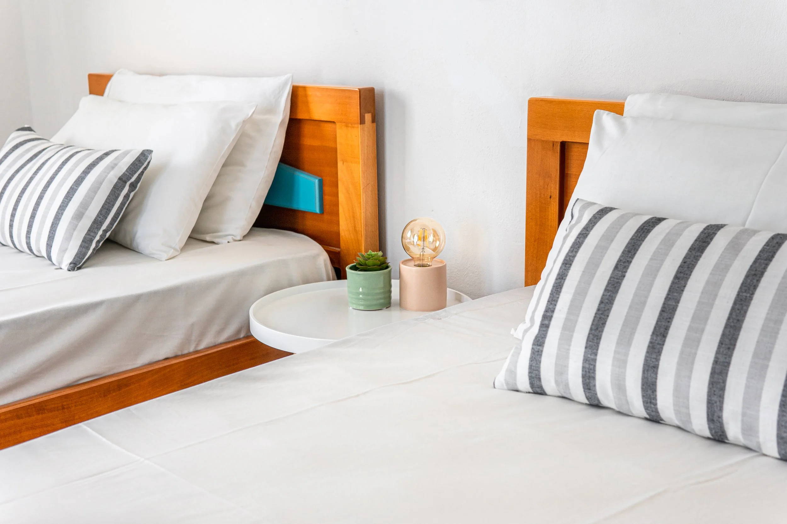 Two beds with wooden headboards, white bedding, and striped pillows in a bright, minimalist bedroom. A small white round table holds two potted plants and a decorative light bulb.