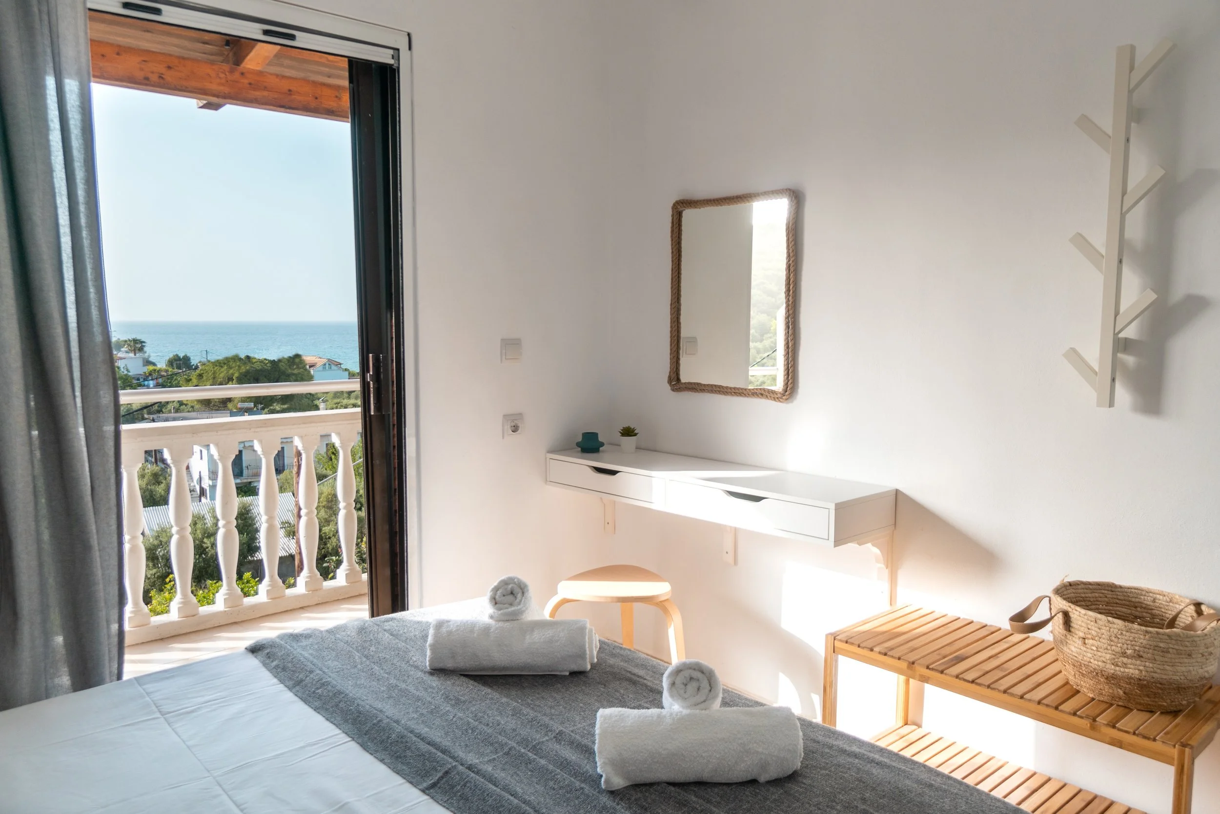 A bedroom with an open balcony door revealing a view of the ocean and houses below. The room has a white wall, a mirror with a rope frame, a small white dresser with a plant and decor items, and a wooden bench. Towels are rolled on the bed.