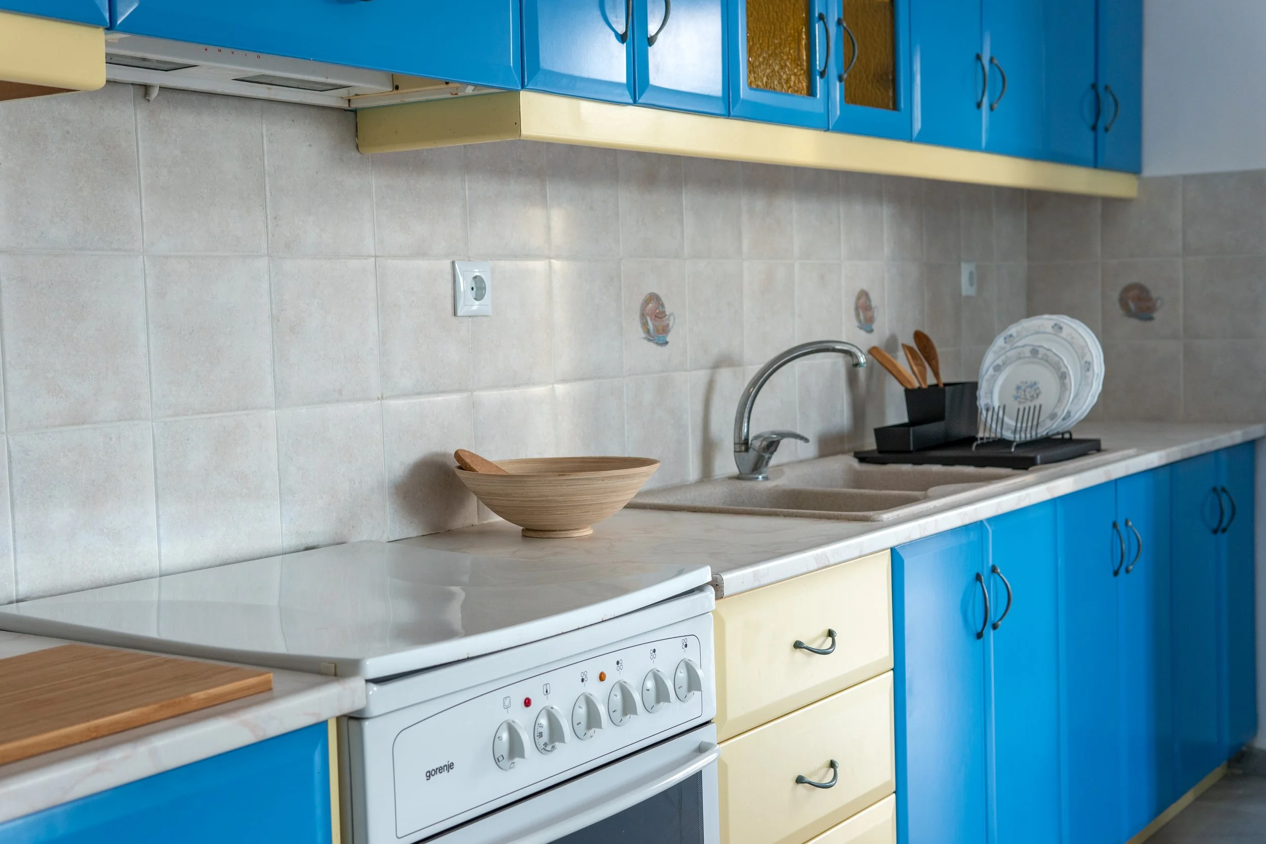 Colorful kitchen with blue and yellow cabinets, tiled backsplash, white stove, and a countertop with a wooden bowl and a dish rack holding plates.