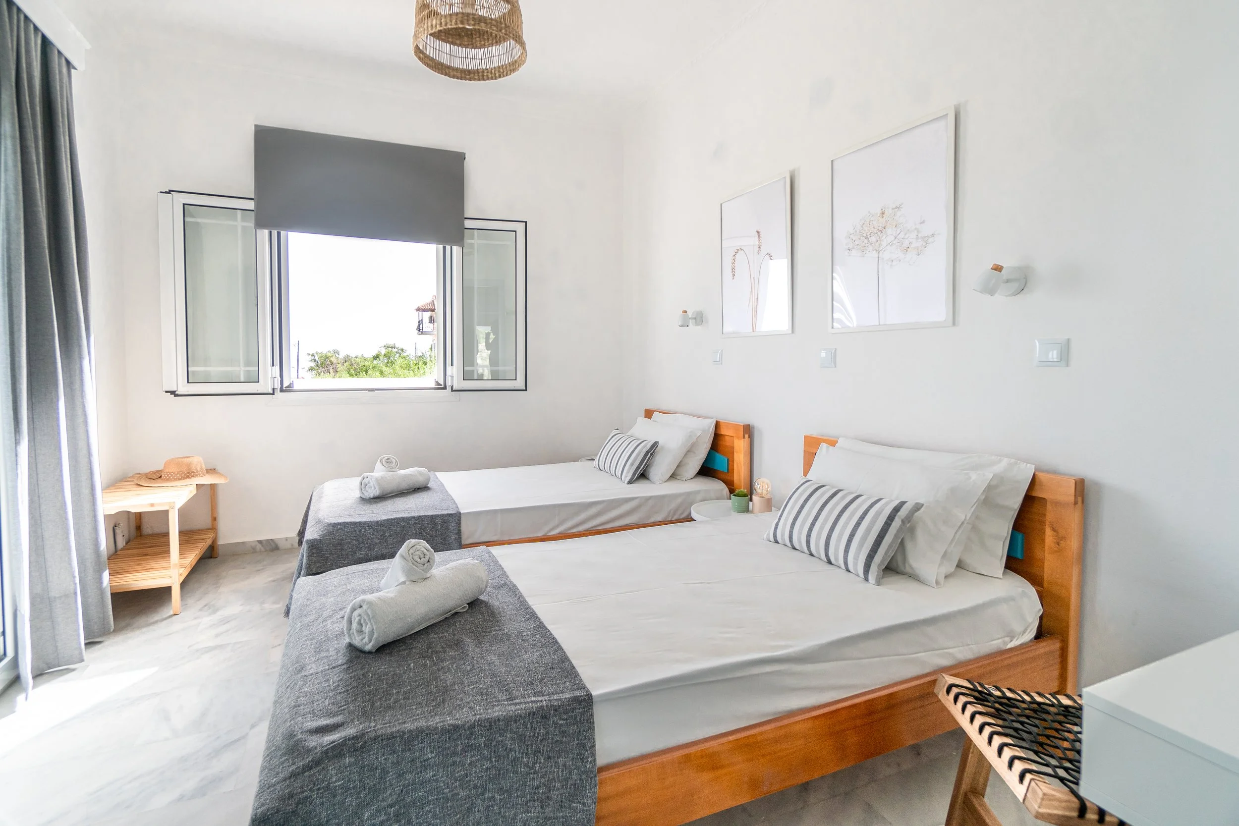 A bright, minimalist bedroom with two single beds, each with white linens and gray-striped pillows, and a window open to a sunny view outside. The room has white walls, a small wooden side table, and decorative framed artwork.