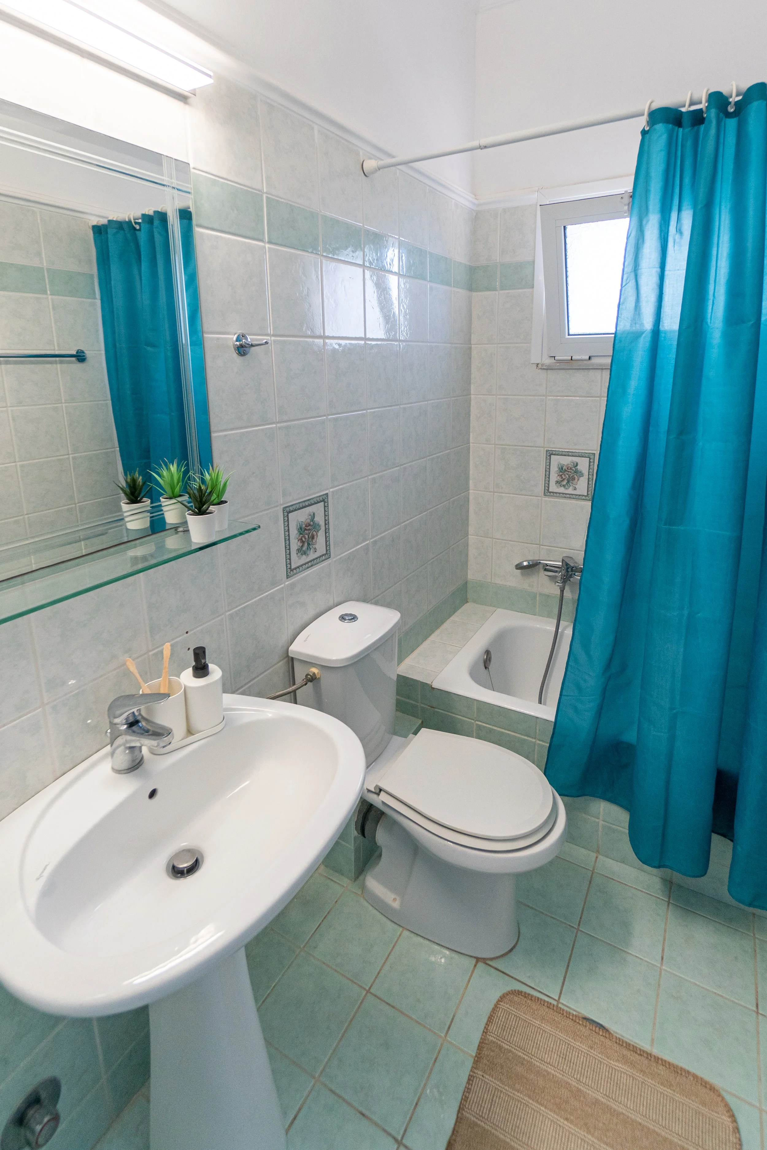 A small bathroom with a white sink, a toilet, and a bathtub with a blue shower curtain. The walls are tiled with light green and white tiles, and there is a small window with a white frame. The bathroom has a mirror above the sink, decorated with small potted plants, and a beige rug on the floor.