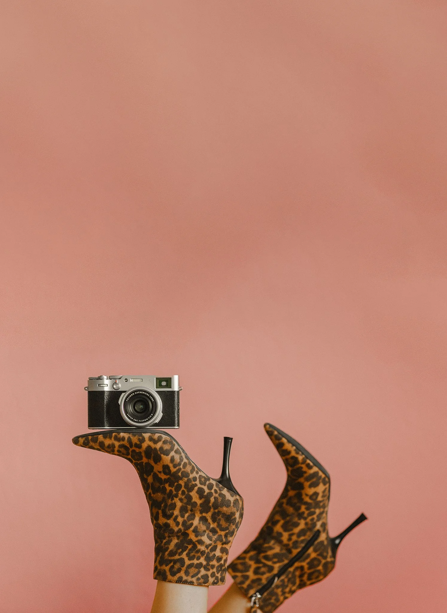 A pair of mannequin legs with leopard print shoes/boots against a pink background, with a vintage camera on top of one leg.