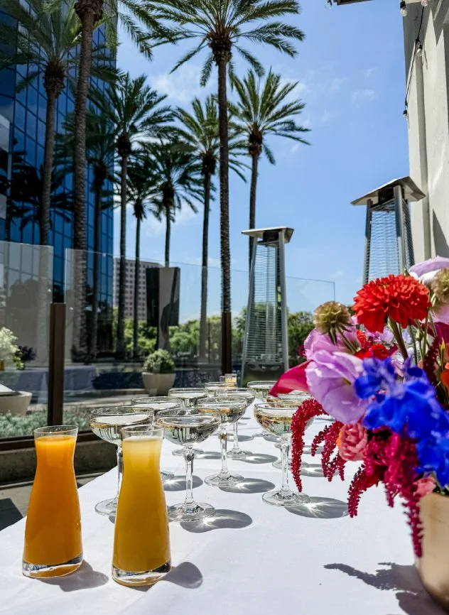 A table set with glasses of orange juice and champagne glasses, decorated with a colorful floral arrangement, outdoors on a patio with tall palm trees and buildings in the background on a sunny day.